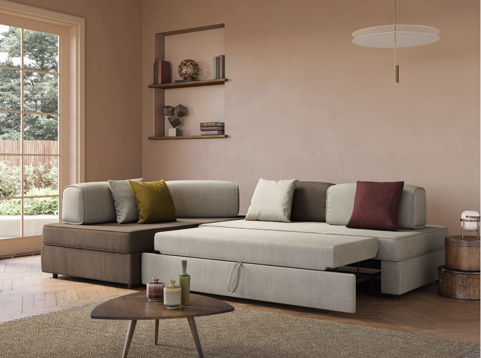 Soggiorno moderno con divano componibile con letto estraibile, cuscini assortiti, tavolino rotondo, mensole a muro con decorazioni, una grande finestra, pareti neutre e una lampada a sospensione circolare.