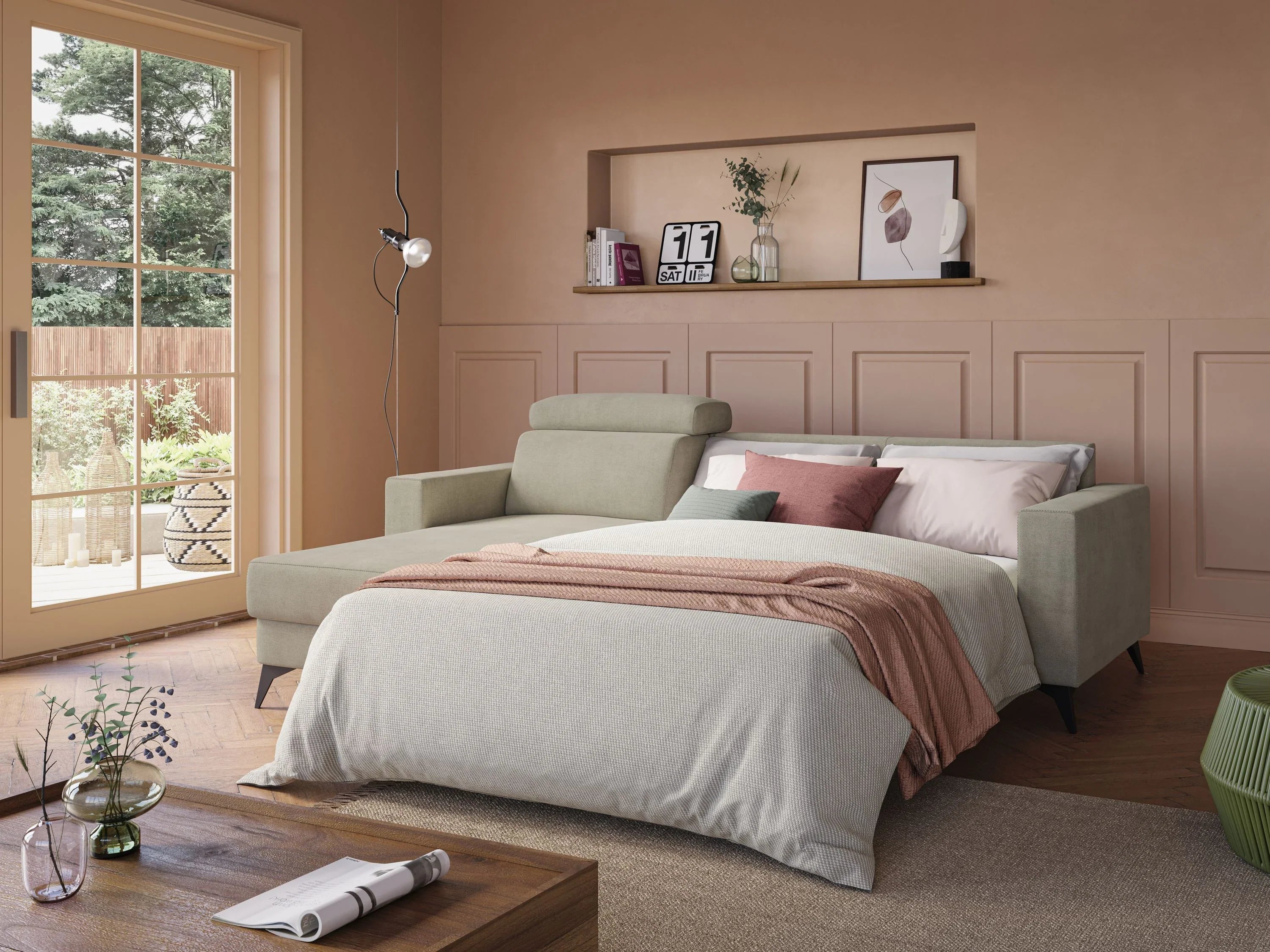 Un divano letto beige con biancheria da letto neutra e dettagli rosa si trova in una stanza accogliente con pareti color cuoio, pavimenti in legno, una mensola con decorazioni e grandi porte a vetri che si affacciano sul giardino.