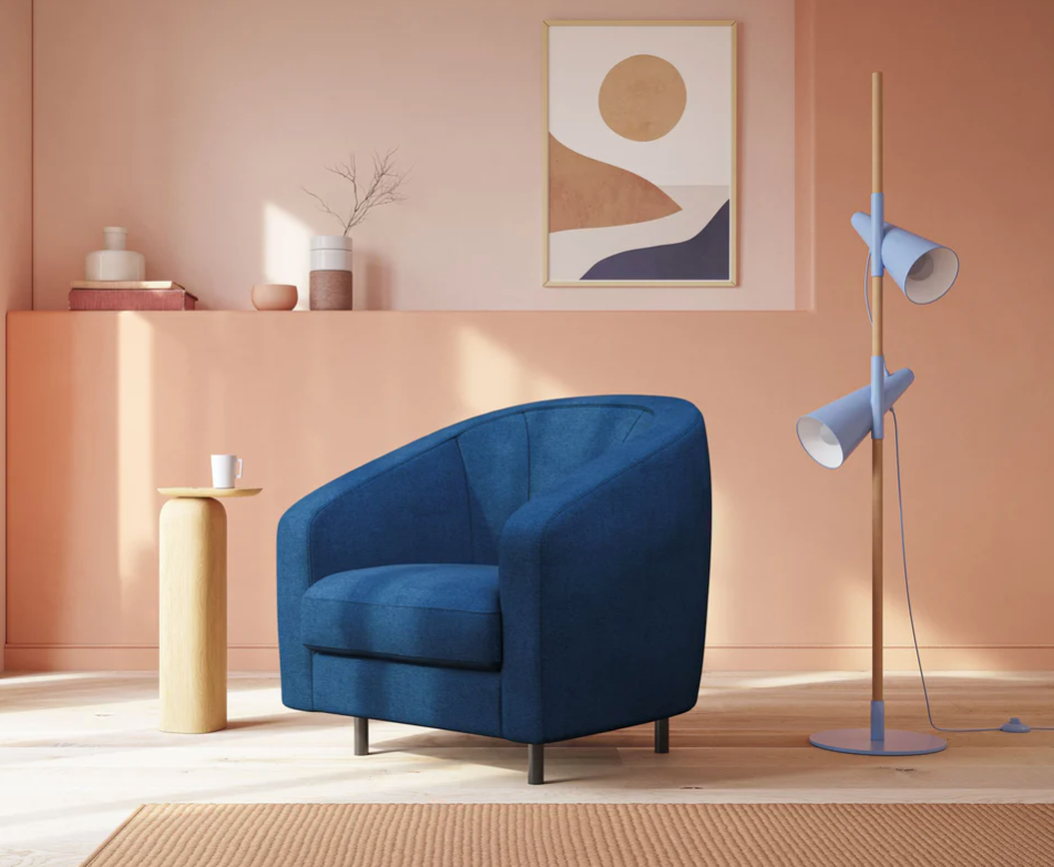 Un soggiorno moderno con una poltrona blu scuro, un tavolino in legno chiaro con una tazza, quadri astratti su una parete color pesca, una lampada da terra blu e decorazioni minimaliste sugli scaffali. La luce del sole entra a fiotti.