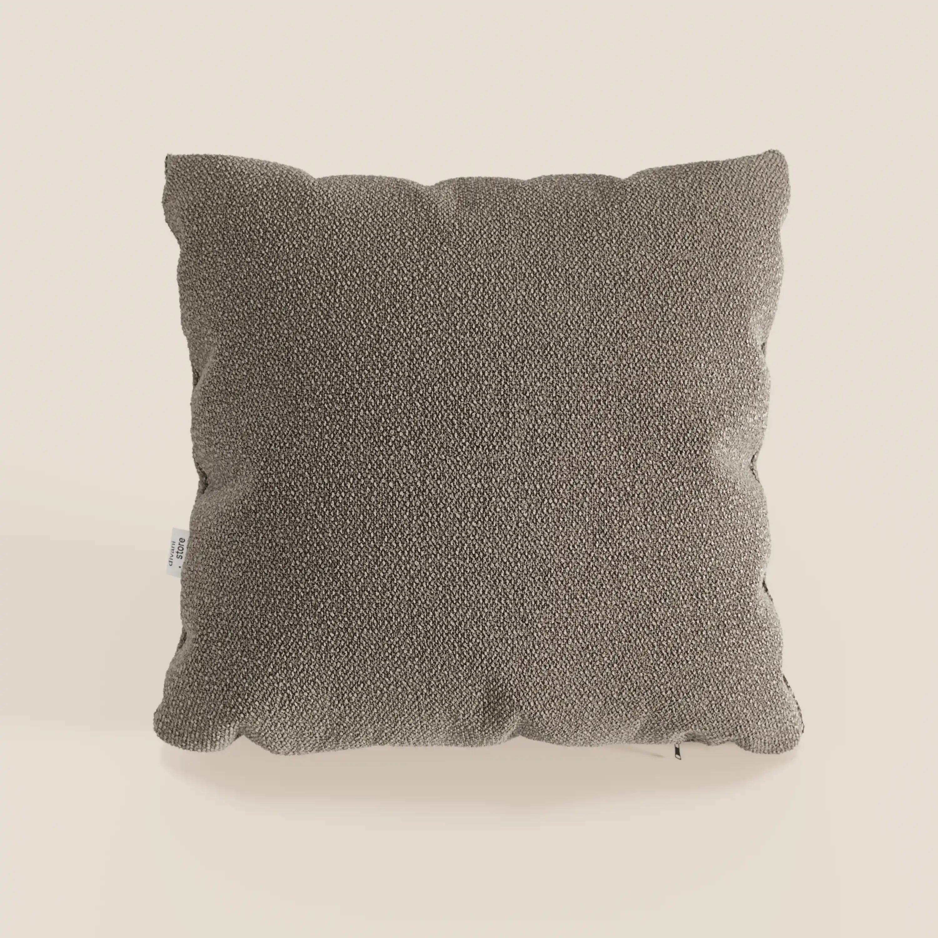 Un cuscino Lambada grigio, quadrato e strutturato, di Divani.Store si trova al centro di uno sfondo bianco sporco. Realizzato in tessuto fresco, antimacchia e impermeabile tipo cotone T33 con tessitura a vista e una piccola etichetta sul bordo sinistro.