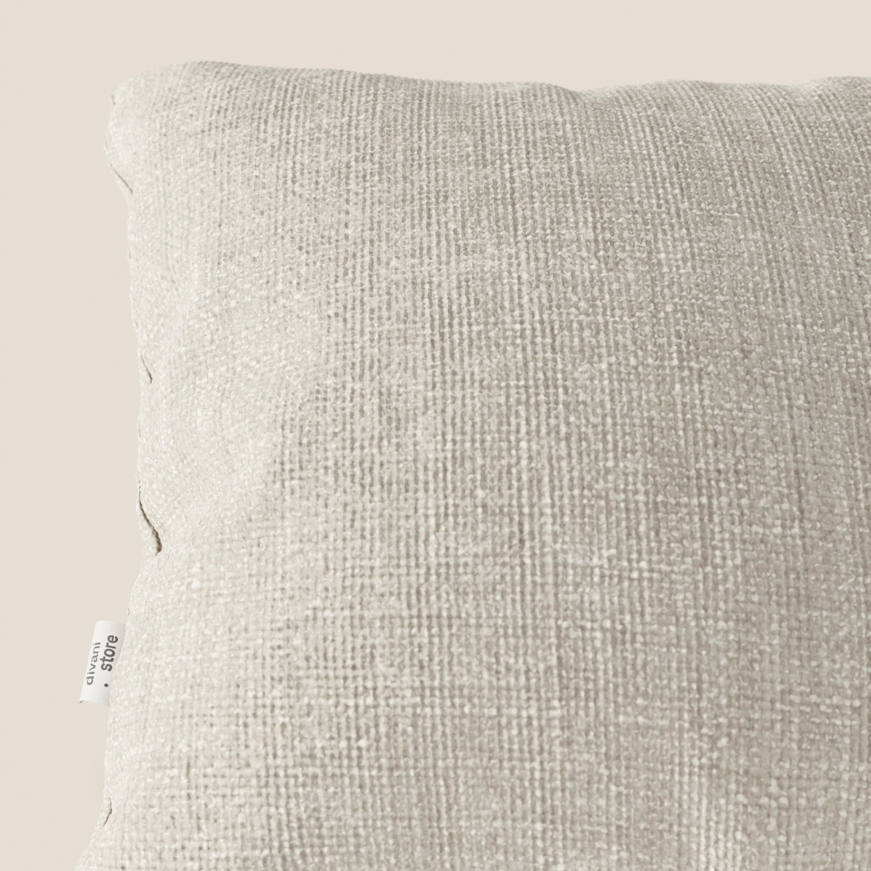Primo piano del cuscino Lyon 45x45 in panna T06 di Divani.Store, realizzato in morbido tessuto impermeabile intrecciato. Su uno sfondo crema chiaro, le cuciture sul bordo e la targhetta bianca ne evidenziano la finitura di pregio.