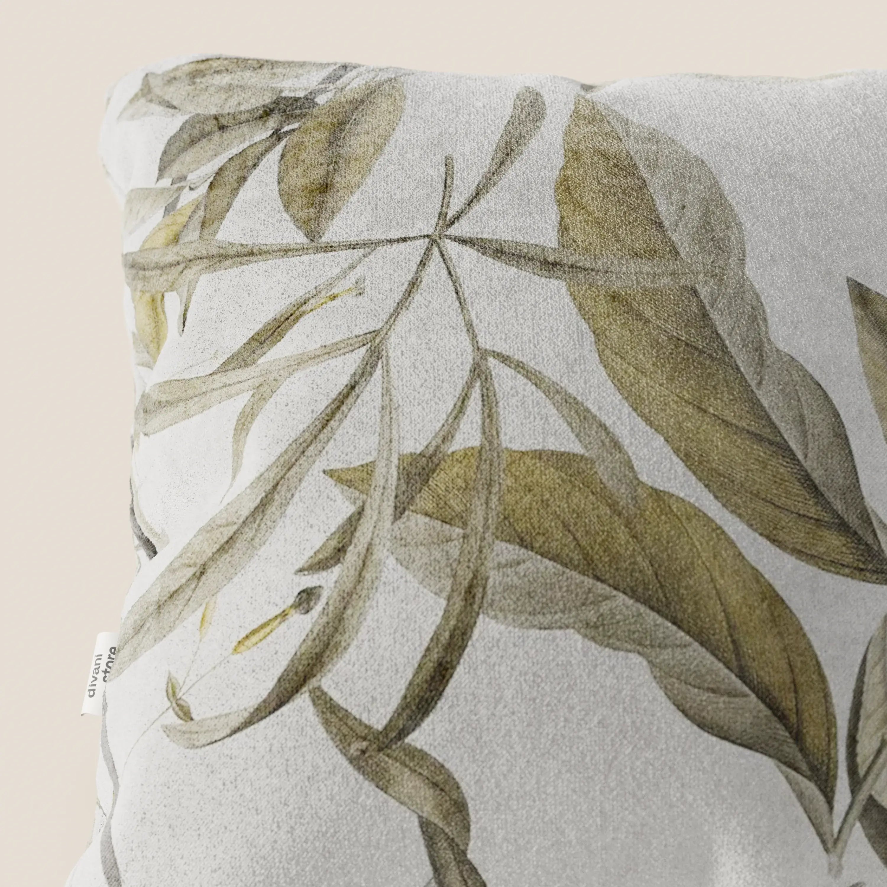 Primo piano del cuscino Lemon elegante a fantasia in velluto impermeabile panna di Divani.Store, che mostra una stampa botanica con foglie verdi e marroni su un tessuto morbido. Una piccola etichetta bianca sottolinea la cucitura su uno sfondo beige chiaro.
