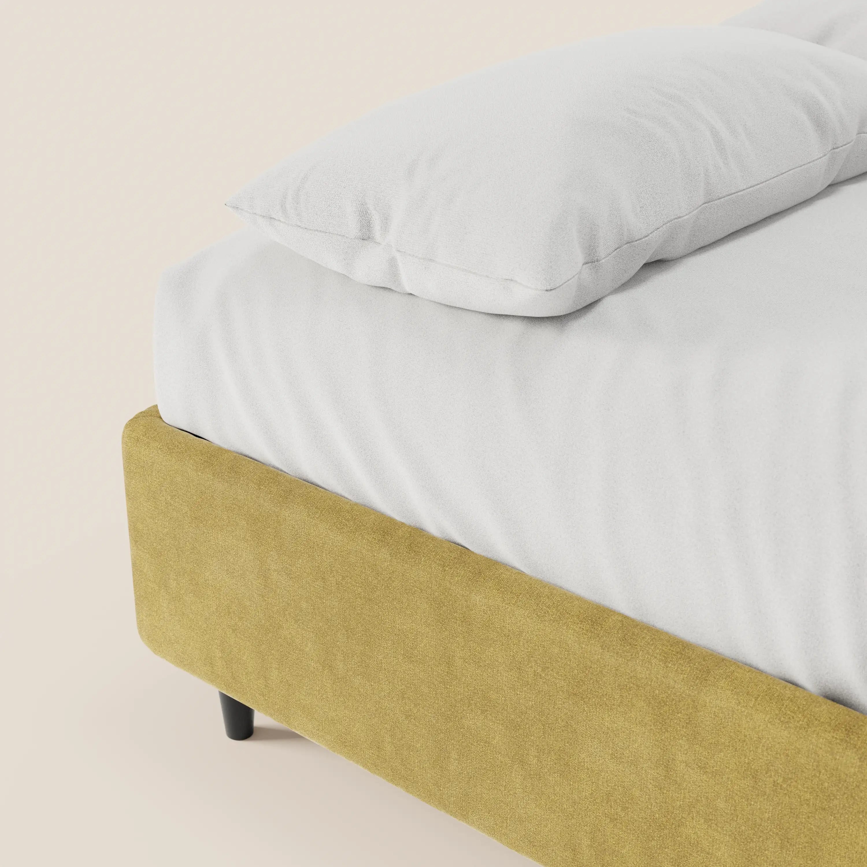 Un primo piano del letto sommier imbottito con contenitore ERON di Letti.store, realizzato in tessuto impermeabile certificato Oeko-Tex, ne mette in risalto il rivestimento giallo senape, le gambe corte nere e il cuscino e il lenzuolo bianchi su uno sfondo chiaro.