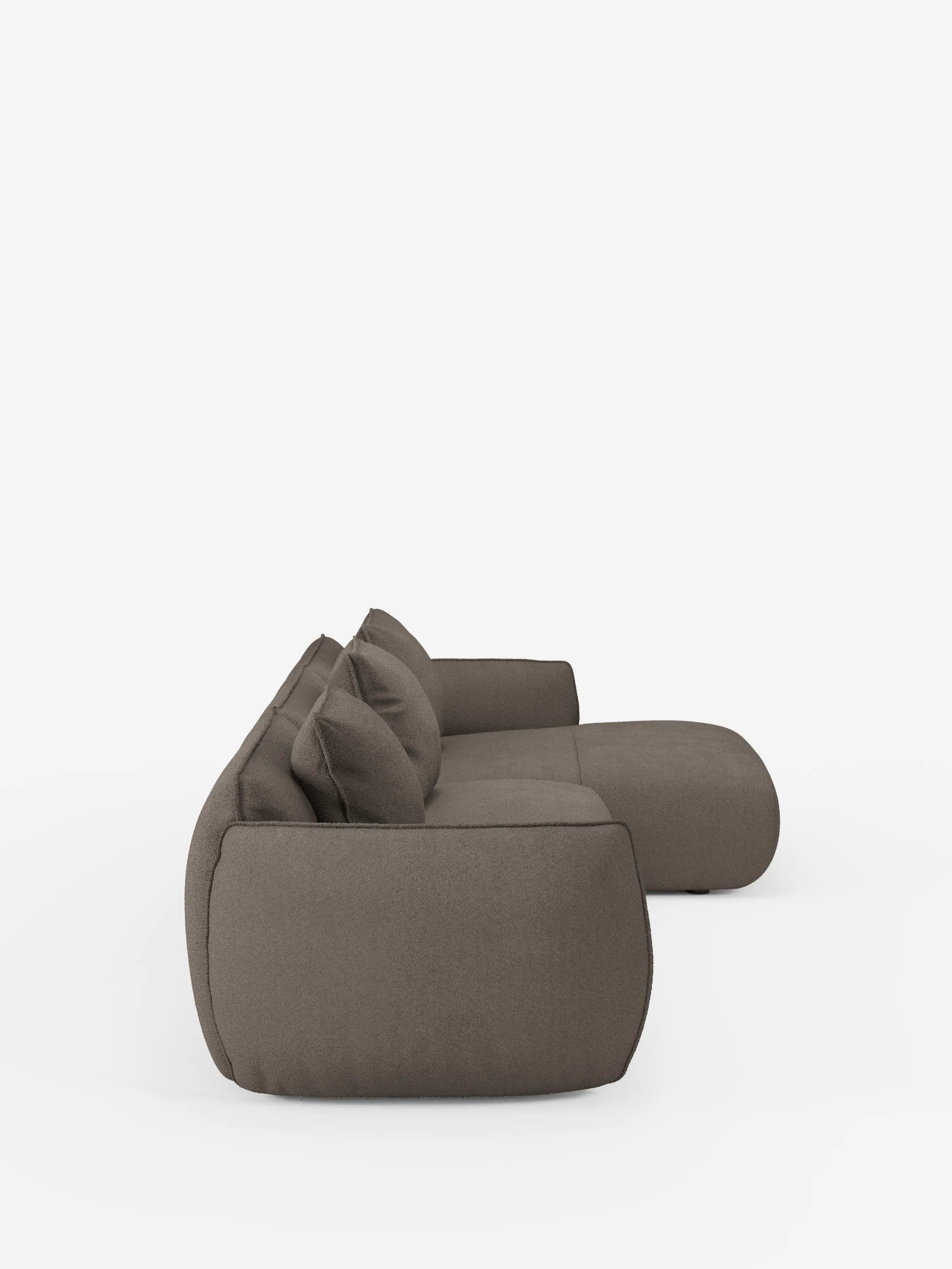 Un moderno divano angolare con penisola componibile Nimbus di colore marrone, firmato Divani.Store, è raffigurato di profilo su uno sfondo bianco. Rivestito in morbido tessuto effetto cotone resistente alle macchie (T33), questo divano modulare dal profilo basso presenta bordi arrotondati, tre grandi cuscini schienali e sedute profonde e avvolgenti, per un look minimalista ma accogliente.
