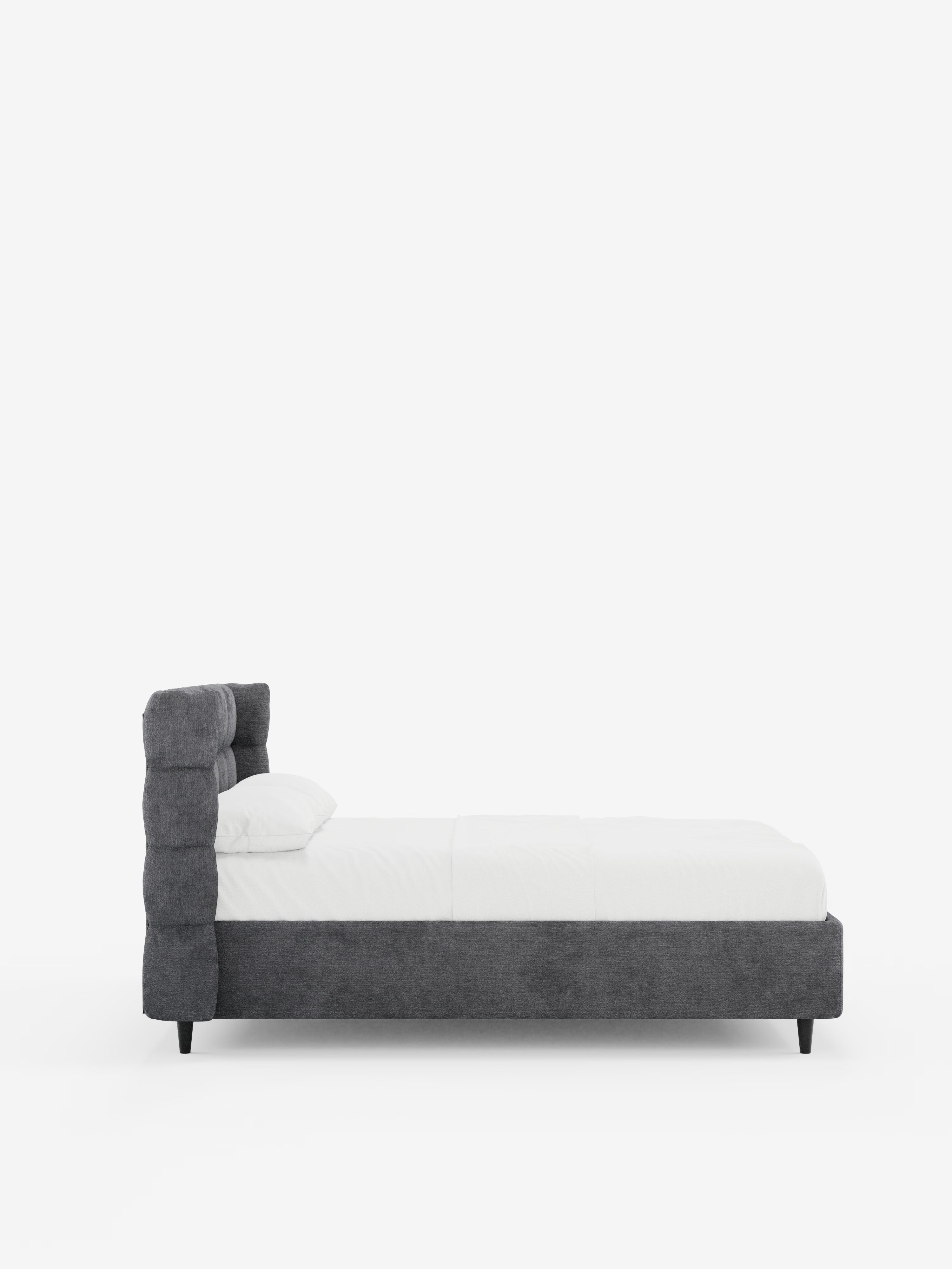 Una vista laterale del letto imbottito RAGNAR con contenitore e testiera capitonné avvolgente di Letti.store, realizzato in un elegante tessuto grigio scuro certificato Oeko-Tex®. Questo letto moderno presenta una testiera alta e trapuntata e poggia su corte gambe nere. Il letto, rifatto con cura, è abbinato a un materasso bianco, biancheria da letto candida e un unico cuscino bianco, il tutto su uno sfondo bianco a tinta unita.