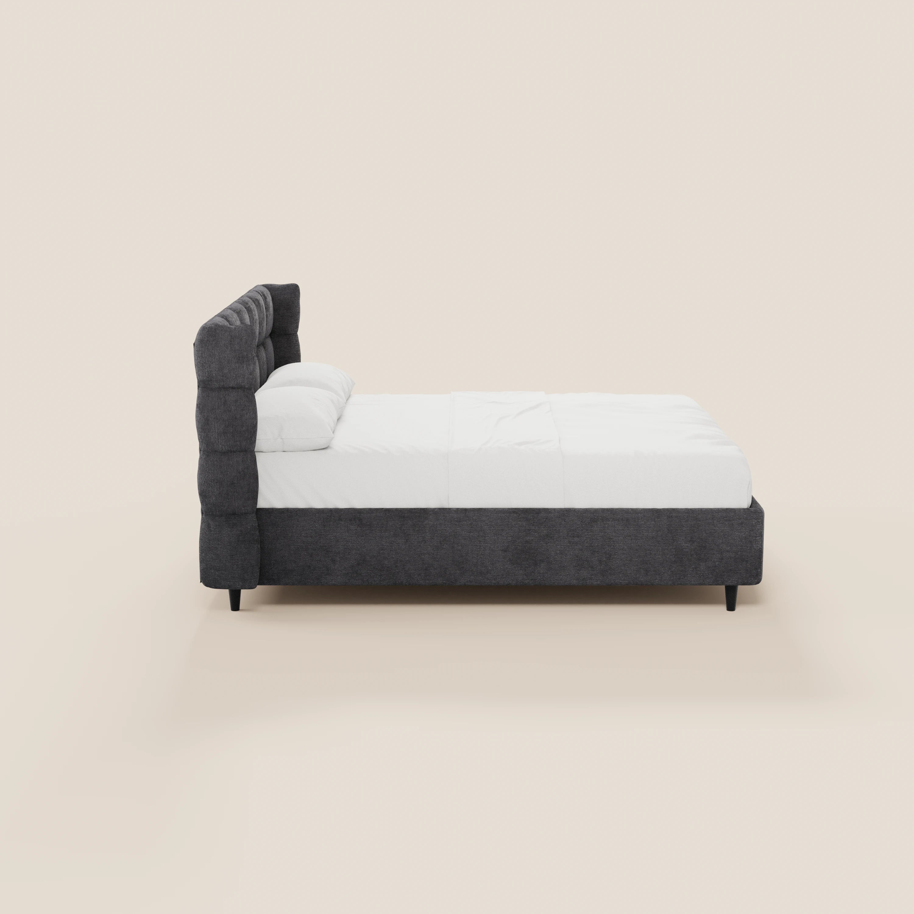 Vista laterale del letto imbottito RAGNAR di Letti.store, caratterizzato da un'alta testiera avvolgente capitonné in tessuto grigio scuro certificato Oeko-Tex®, biancheria da letto bianca, due cuscini, gambe corte nere e un design minimalista su uno sfondo beige chiaro.