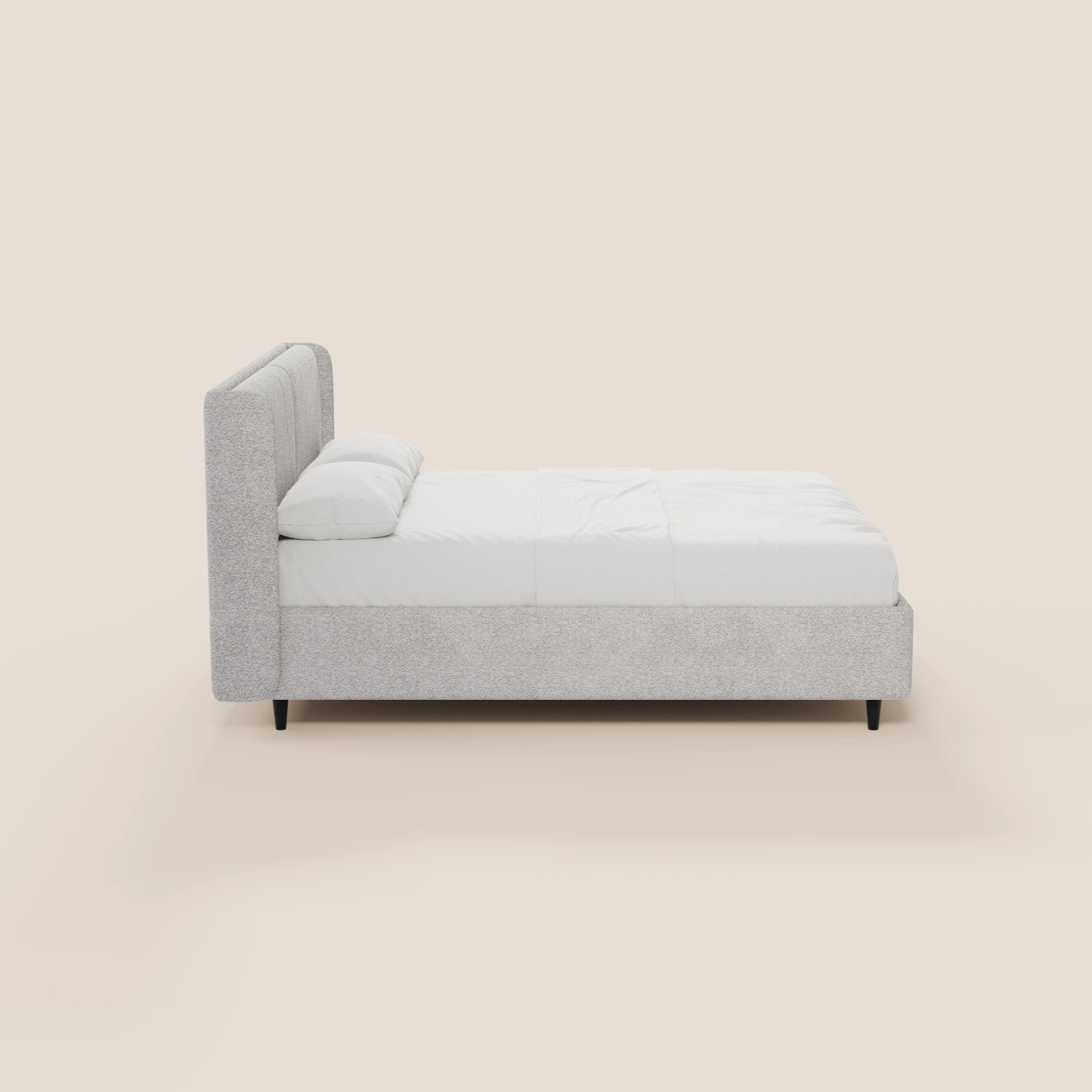 Vista laterale del letto di design VALKIRIA di Letti.store, caratterizzato da una testiera alta e imbottita, 2 cuscini imbottiti, tessuto Oeko-Tex® in grigio chiaro, biancheria da letto bianca, gambe corte nere e un vano contenitore su uno sfondo bianco sporco.