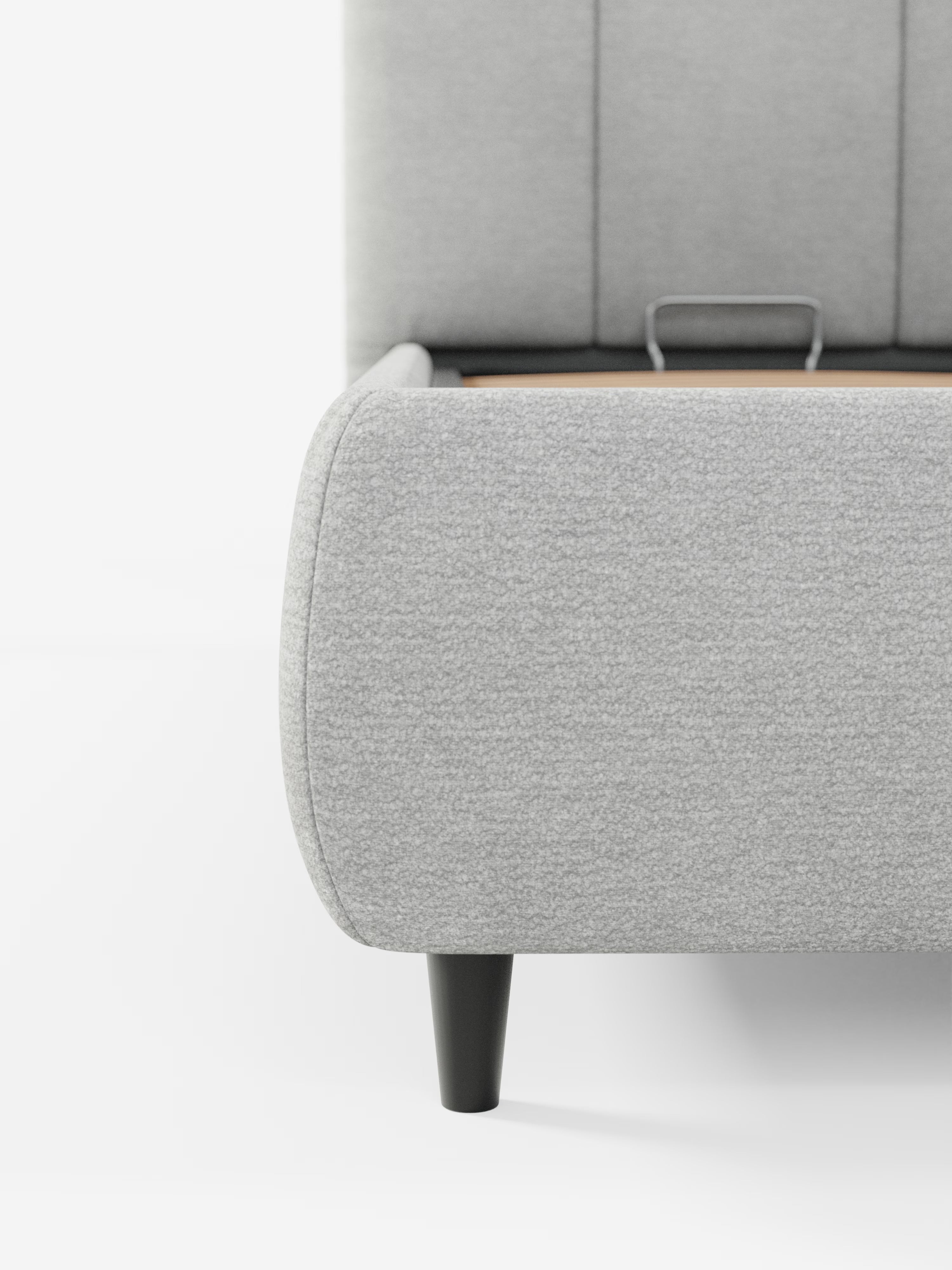 Primo piano del letto imbottito ARKAM con vano contenitore e testiera a lavorazione verticale di Letti.store, caratterizzato da un moderno tessuto grigio chiaro certificato Oeko-Tex® e da gambe affusolate nere. L’immagine mostra parte delle doghe in legno del letto e una piccola maniglia in metallo all’interno della struttura, mettendo in risalto la sua pratica funzione di contenitore (letto contenitore). Su uno sfondo bianco a tinta unita, la foto sottolinea la morbida consistenza del letto e le sue linee