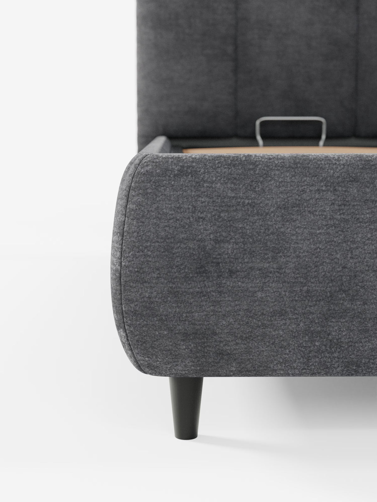 Primo piano del letto imbottito ARKAM con contenitore e testiera a lavorazione verticale di Letti.store, che mette in risalto il rivestimento in tessuto grigio scuro certificato Oeko-Tex®, gli angoli arrotondati con maniglia integrata tipici del design pratico dei letti contenitore e le eleganti gambe affusolate nere. Lo sfondo bianco a tinta unita mette in risalto lo stile shabby chic e la trama raffinata di questo letto moderno.