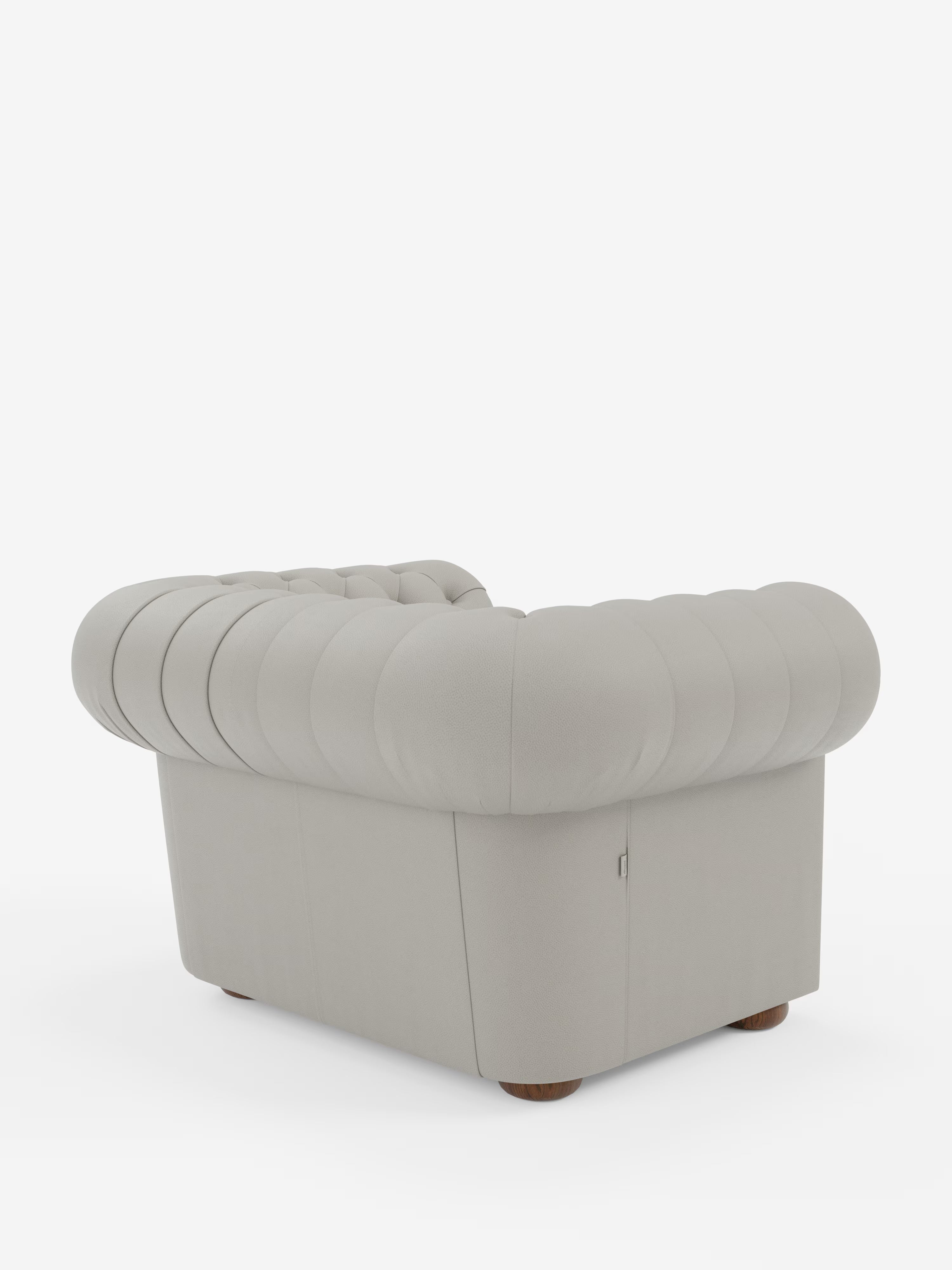 Una lussuosa poltrona Chester in vera pelle spessorata con capitonné artigianale di Divani.Store: questa poltrona grigio chiaro presenta uno schienale arrotondato e trapuntato e braccioli con un’elegante lavorazione capitonné. Il morbido rivestimento in pelle è piacevole al tatto, mentre le gambe corte e rotonde in legno garantiscono un solido sostegno. La foto mette in risalto questa raffinata poltrona in pelle, ripresa da un’angolazione posteriore sinistra su uno sfondo bianco a tinta unita.