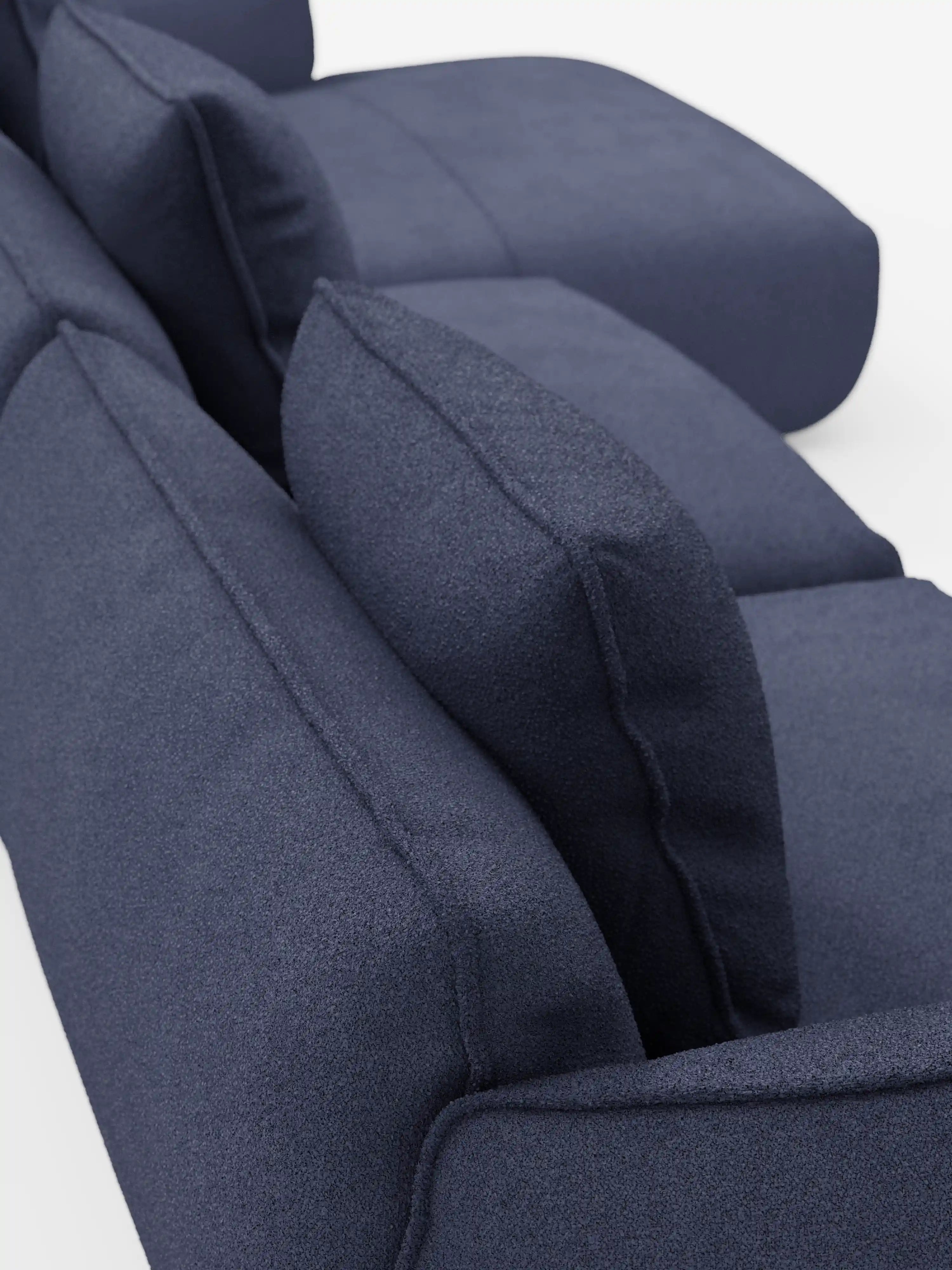 Primo piano, vista angolare del divano angolare con penisola componibile Nimbus di Divani.Store, rivestito in tessuto T33 smacchiabile blu scuro con una morbida trama effetto cotone. L’immagine mette in risalto i cuscini e i braccioli spessi e testurizzati su uno sfondo bianco, concentrandosi sul design morbido e strutturato e sul tessuto ricco di dettagli dei cuscini dello schienale e della seduta.