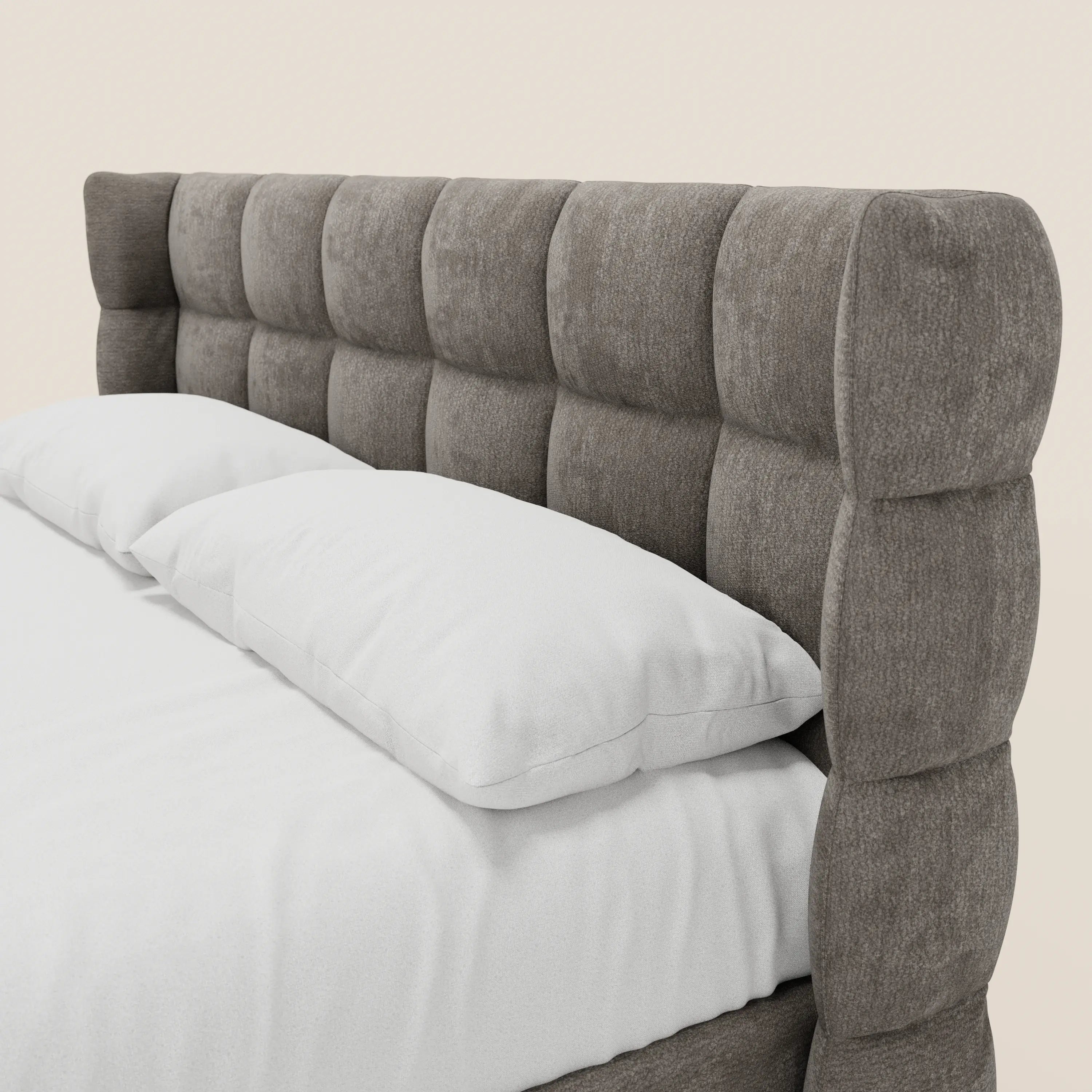 Un primo piano del letto imbottito RAGNAR con contenitore e testiera capitonné avvolgente di Letti.store, realizzato in elegante tessuto Oeko-Tex®, abbinato a cuscini e lenzuola bianchi su uno sfondo beige chiaro.