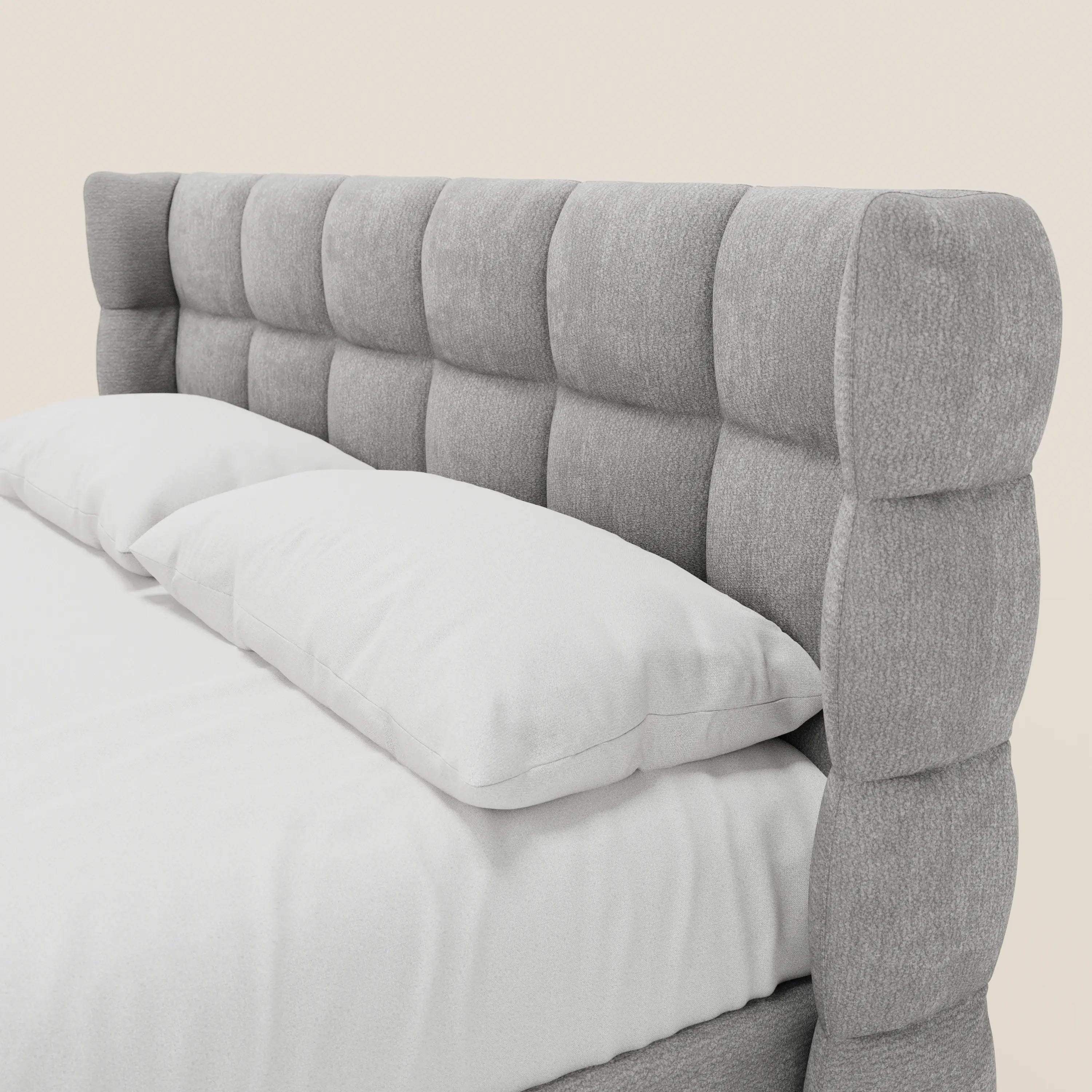 Primo piano del letto imbottito RAGNAR con contenitore e testiera capitonné avvolgente di Letti.store, realizzato in elegante tessuto grigio certificato Oeko-Tex®, completato da un lenzuolo con angoli e due cuscini bianchi su sfondo beige chiaro.