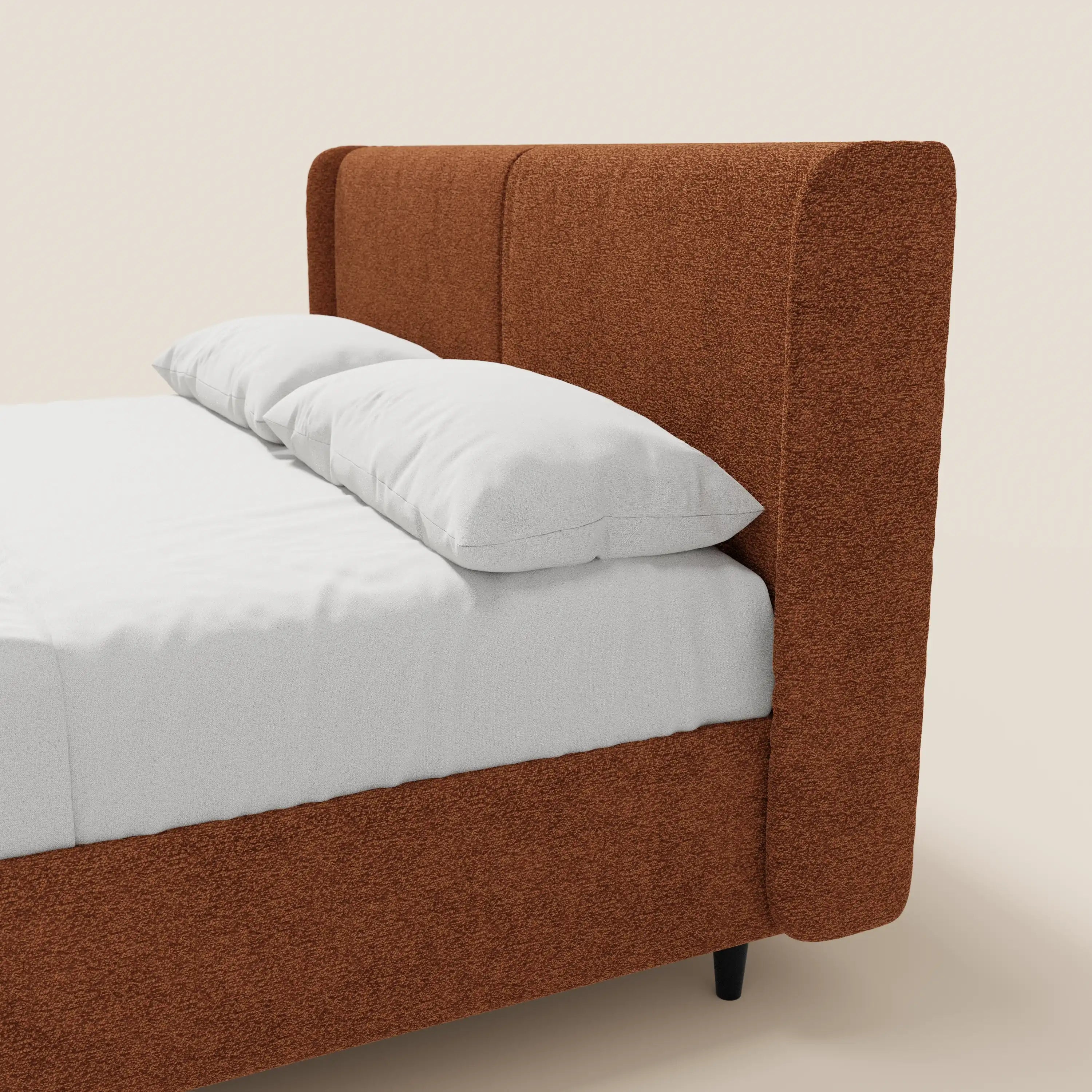 Il letto di design VALKIRIA di Letti.store presenta una struttura rivestita in tessuto Oeko-Tex® color ruggine e dalla trama ruvida, una testiera avvolgente con 2 cuscini imbottiti, biancheria da letto di un bianco brillante, gambe nere e uno sfondo beige chiaro che conferiscono al letto un look minimalista.