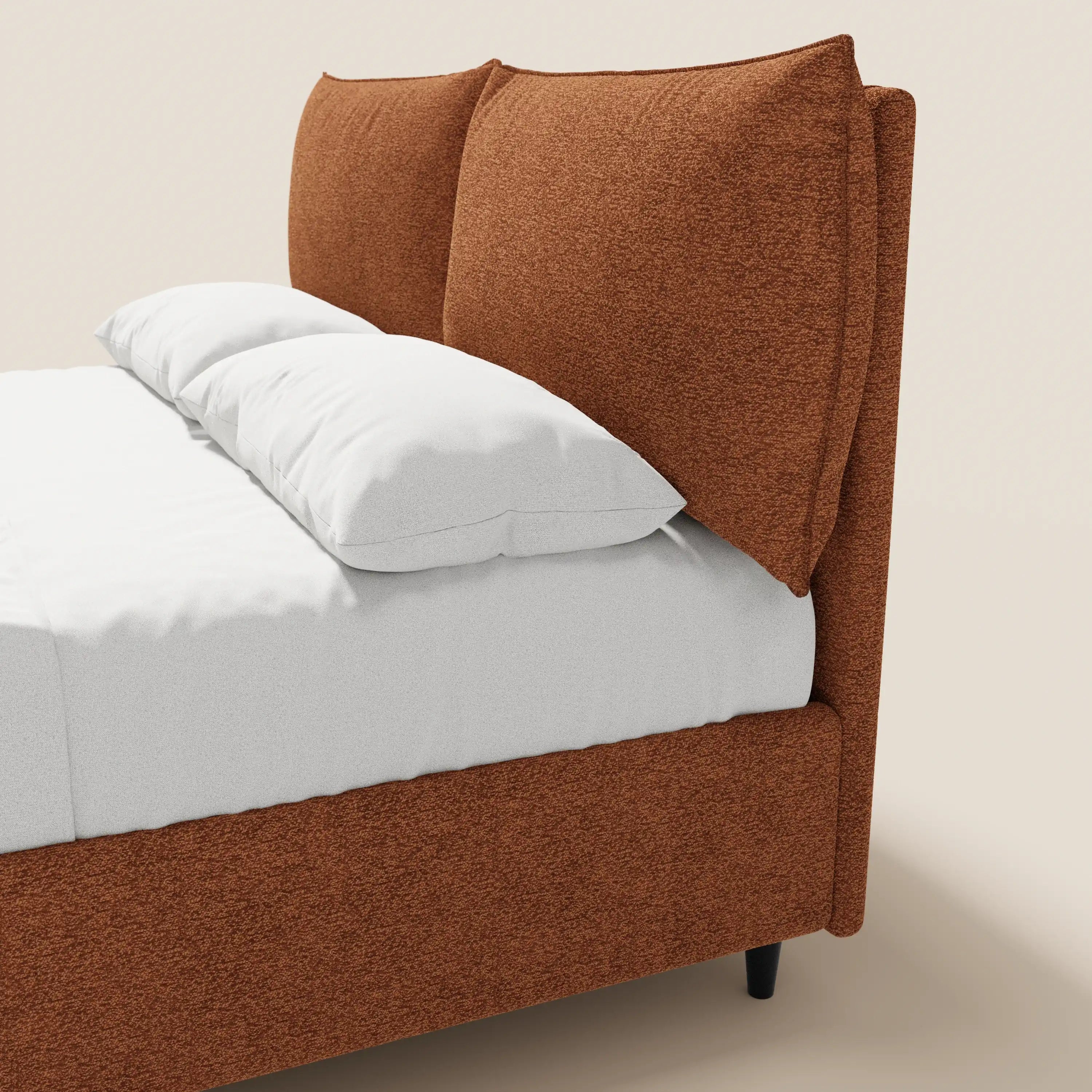 Un primo piano di ZAYRON di Letti.store, un letto imbottito in stile moderno con contenitore e morbidi cuscini regolabili in tessuto certificato Oeko-Tex®, presentato in tessuto color ruggine con lenzuola bianchissime e gambe corte nere su uno sfondo chiaro.
