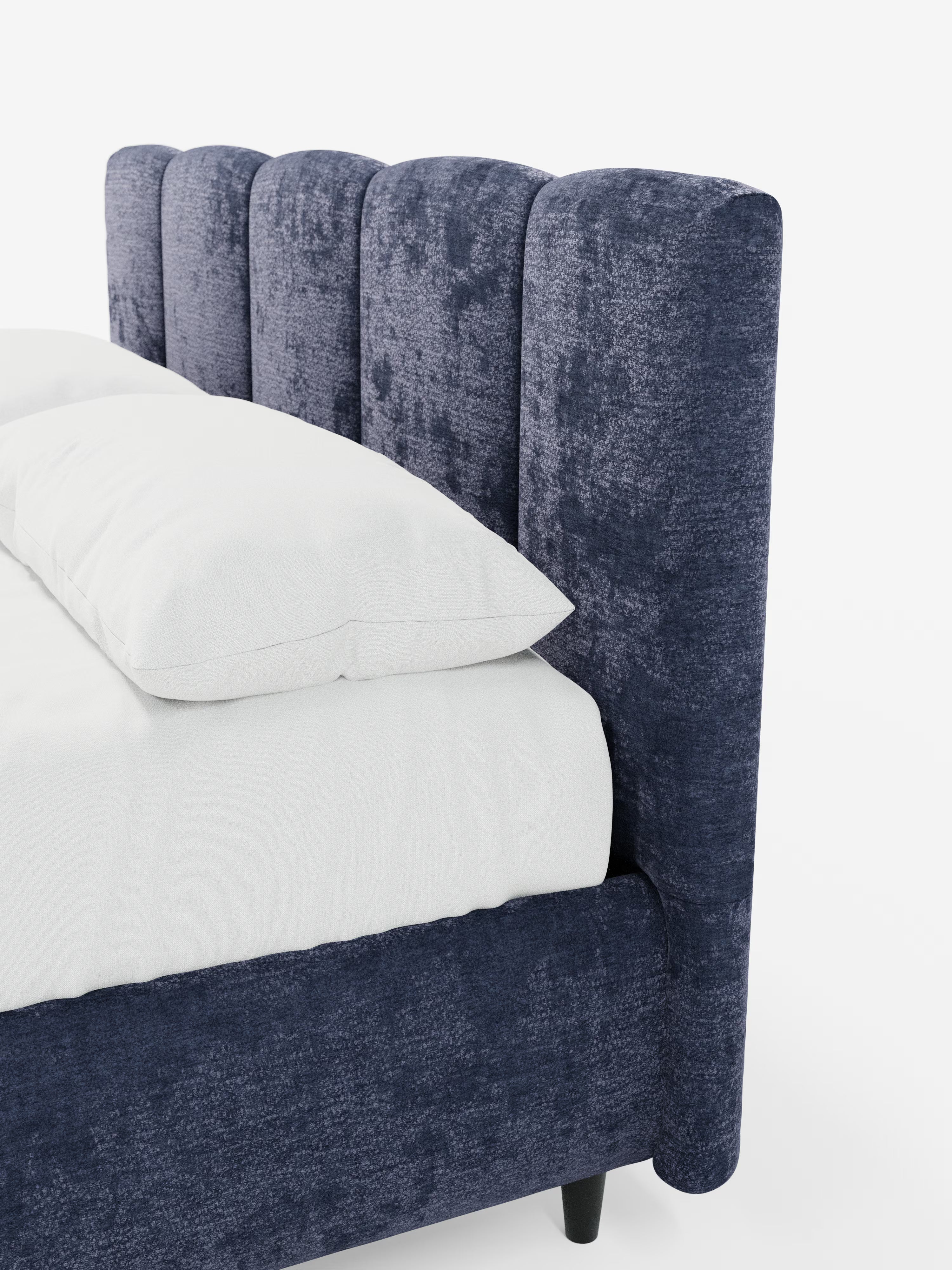 Primo piano di un angolo del letto che mette in risalto il modello ARKAM di Letti.store: un letto imbottito con vano contenitore e testiera a lavorazione verticale. Questa testiera alta e morbida, in blu navy, presenta cuciture a canalina verticali e incarna lo stile shabby chic grazie al tessuto certificato Oeko-Tex®. Il letto, rivestito con biancheria da letto bianca e con il materasso a vista, risalta sullo sfondo bianco liscio, mettendo in risalto le ricche trame e il colore intenso di questo elegante
