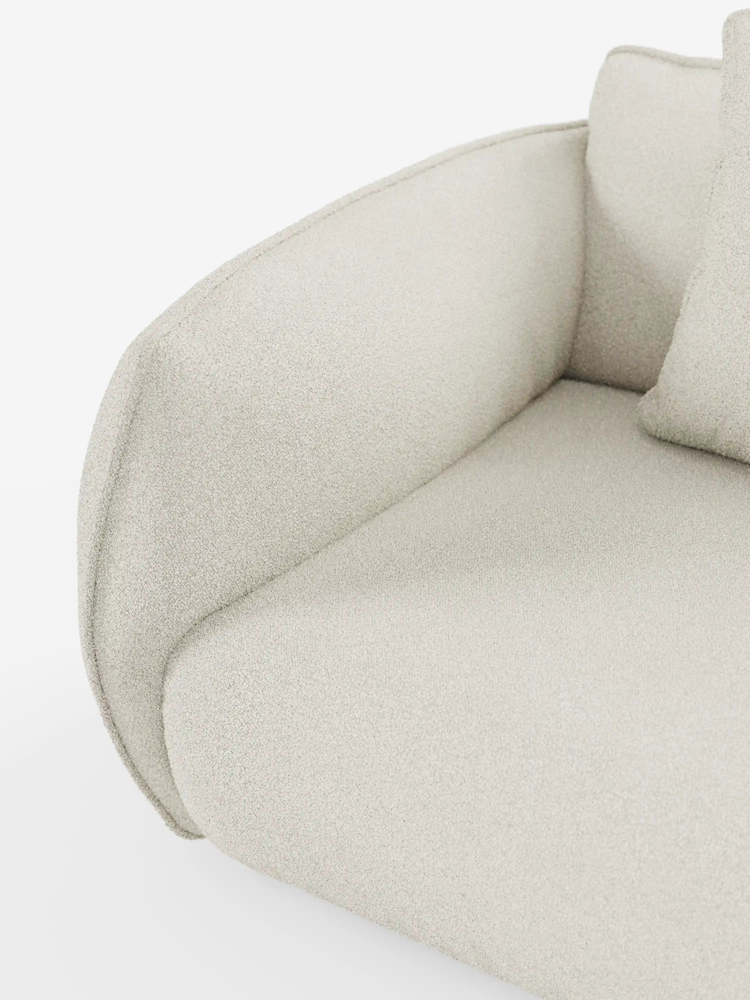 Primo piano del divano angolare con penisola componibile Nimbus di Divani.Store, che mette in risalto i braccioli e la seduta curvi e imbottiti, rivestiti in tessuto T33 effetto cotone beige chiaro, testurizzato e resistente alle macchie. È visibile una parte di un cuscino coordinato, che mette in risalto il design minimalista del divano, caratterizzato da linee pulite e da un aspetto morbido e accogliente.