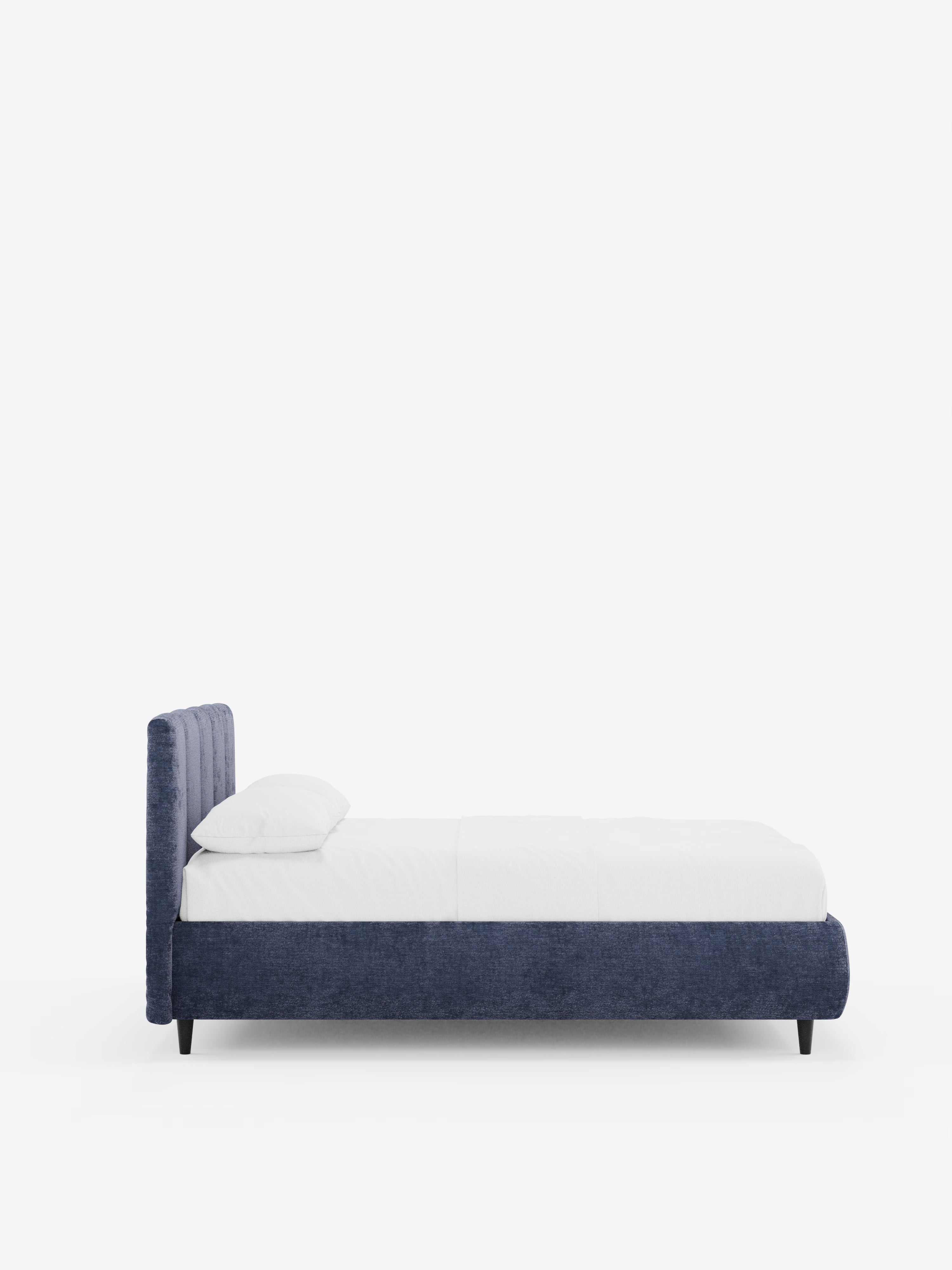 Vista laterale del letto imbottito ARKAM con contenitore e testiera a cuciture verticali di Letti.store, caratterizzato da una struttura in tessuto blu certificato Oeko-Tex®, gambe basse nere, testiera imbottita con cuciture verticali e biancheria da letto bianca con un unico cuscino su uno sfondo bianco a tinta unita.