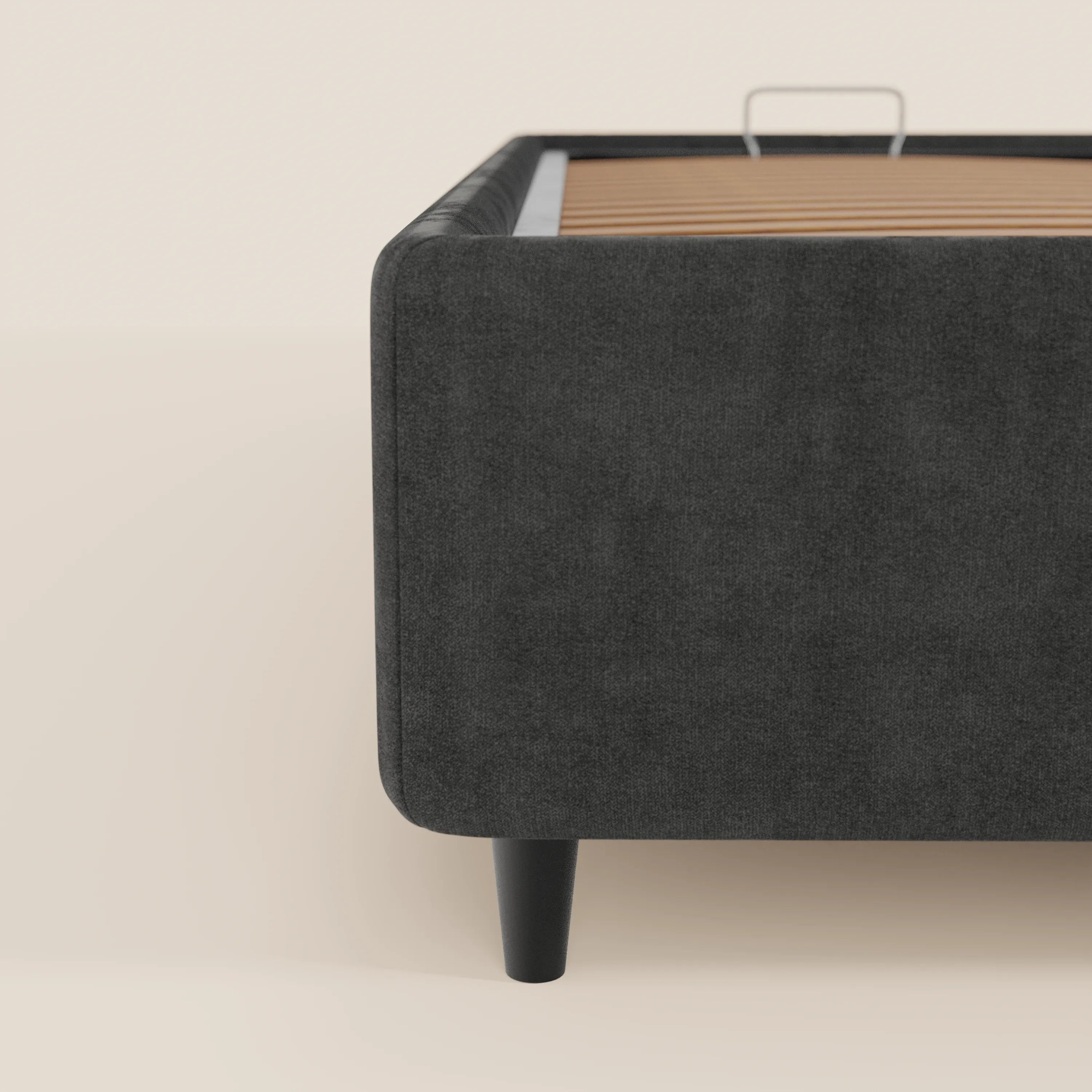Primo piano del letto sommier imbottito con contenitore ERON di Letti.store, realizzato in tessuto impermeabile certificato Oeko-Tex color grigio scuro, con angoli arrotondati e gambe cilindriche nere. Le doghe in legno sono a vista; lo sfondo bianco sporco ne esalta le linee pulite.