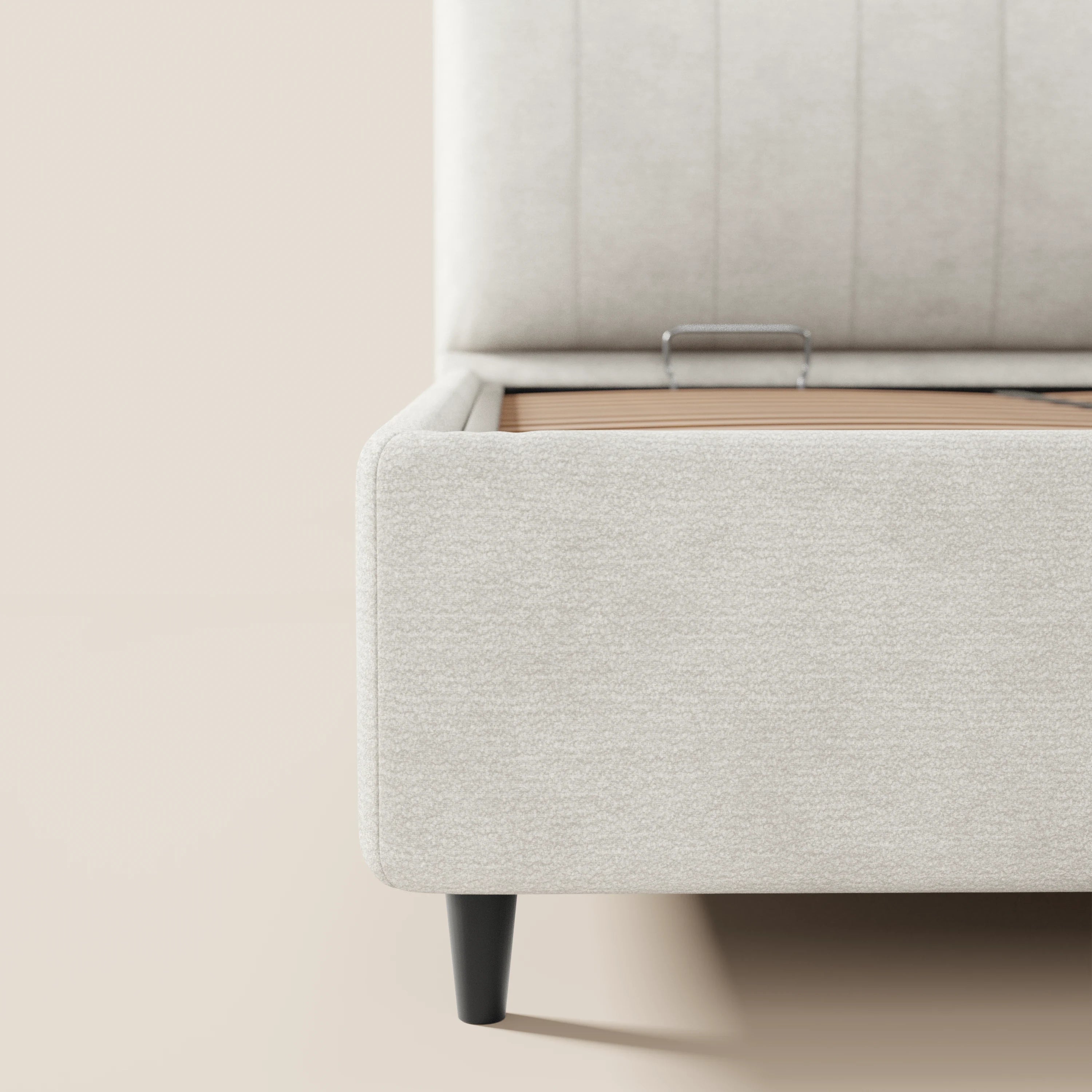 Primo piano del letto imbottito ONYX con contenitore di Letti.store, realizzato in tessuto beige chiaro certificato Oeko-Tex®, caratterizzato da un'alta testiera verticale; il vano contenitore è a vista e presenta interni in legno color cuoio. Questo letto in stile moderno poggia su corte gambe cilindriche nere.
