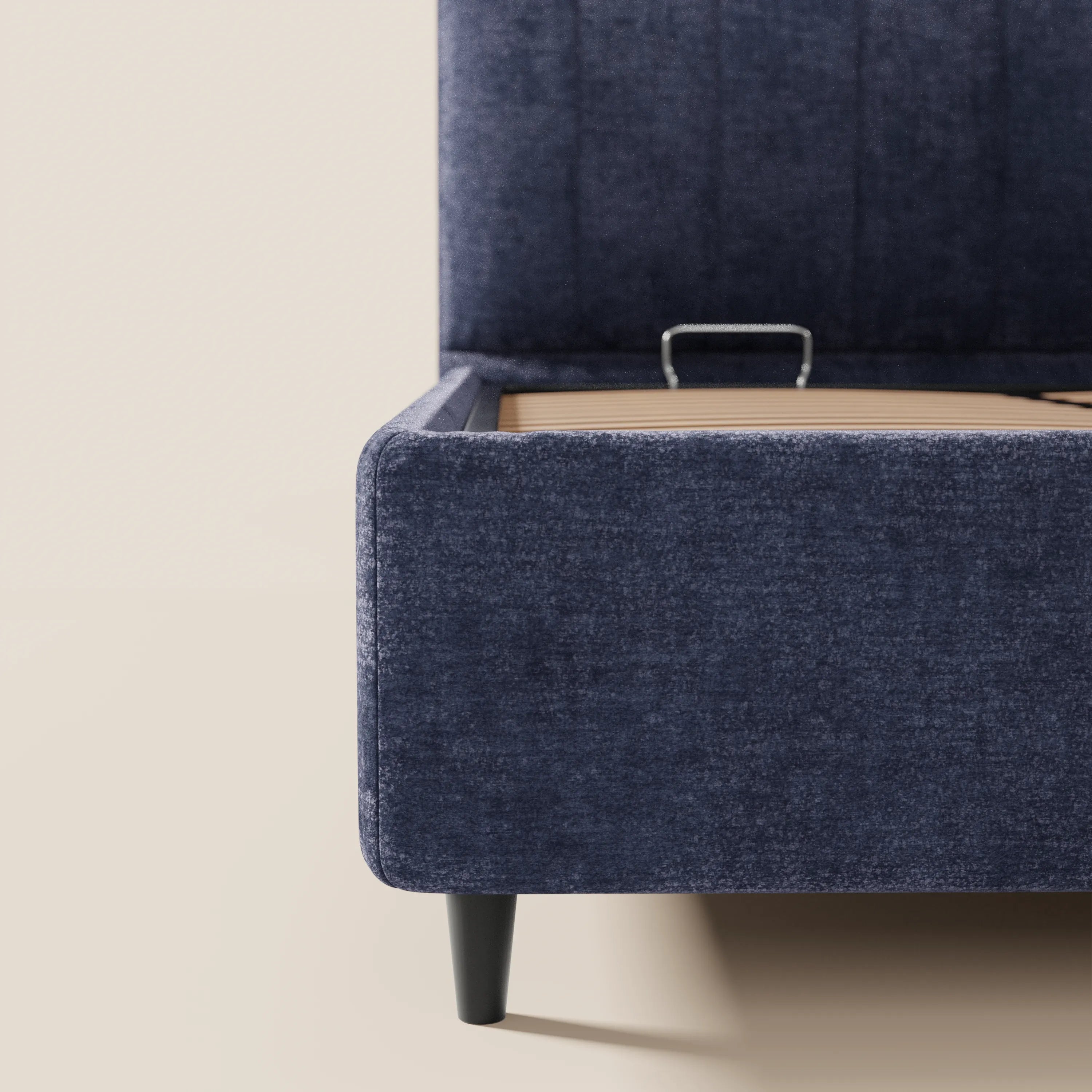 Un primo piano mostra un angolo del letto imbottito con contenitore ONYX di Letti.store, caratterizzato da una base sollevabile con maniglia in metallo, gambe corte nere e una testiera verticale in tessuto certificato Oeko-Tex® su uno sfondo beige chiaro.