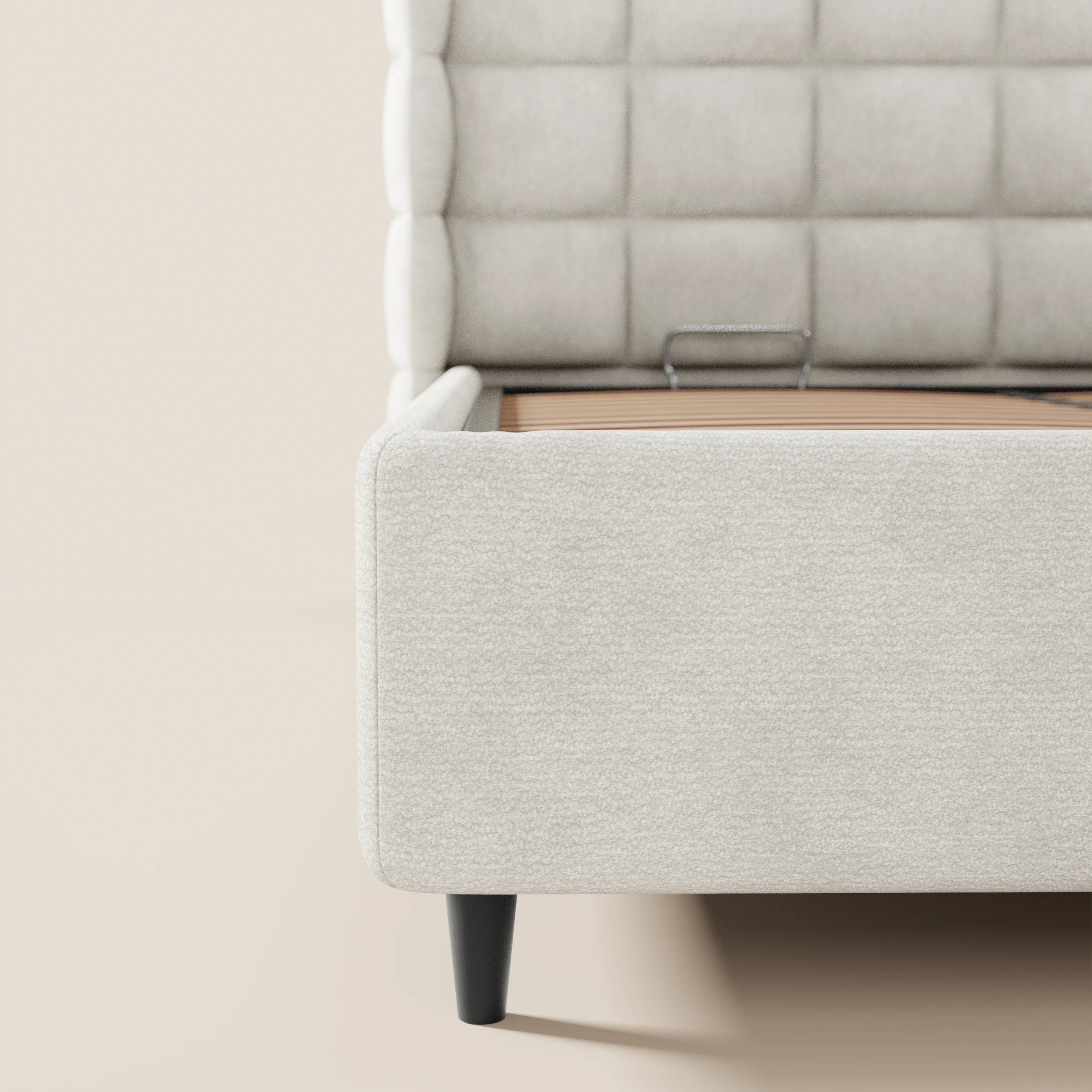 Primo piano del letto imbottito con contenitore RAGNAR di Letti.store, realizzato in tessuto beige chiaro certificato Oeko-Tex® con testiera capitonné; nell’immagine è visibile solo il lato sinistro, che mette in risalto le doghe in legno e le gambe nere angolate su uno sfondo neutro.