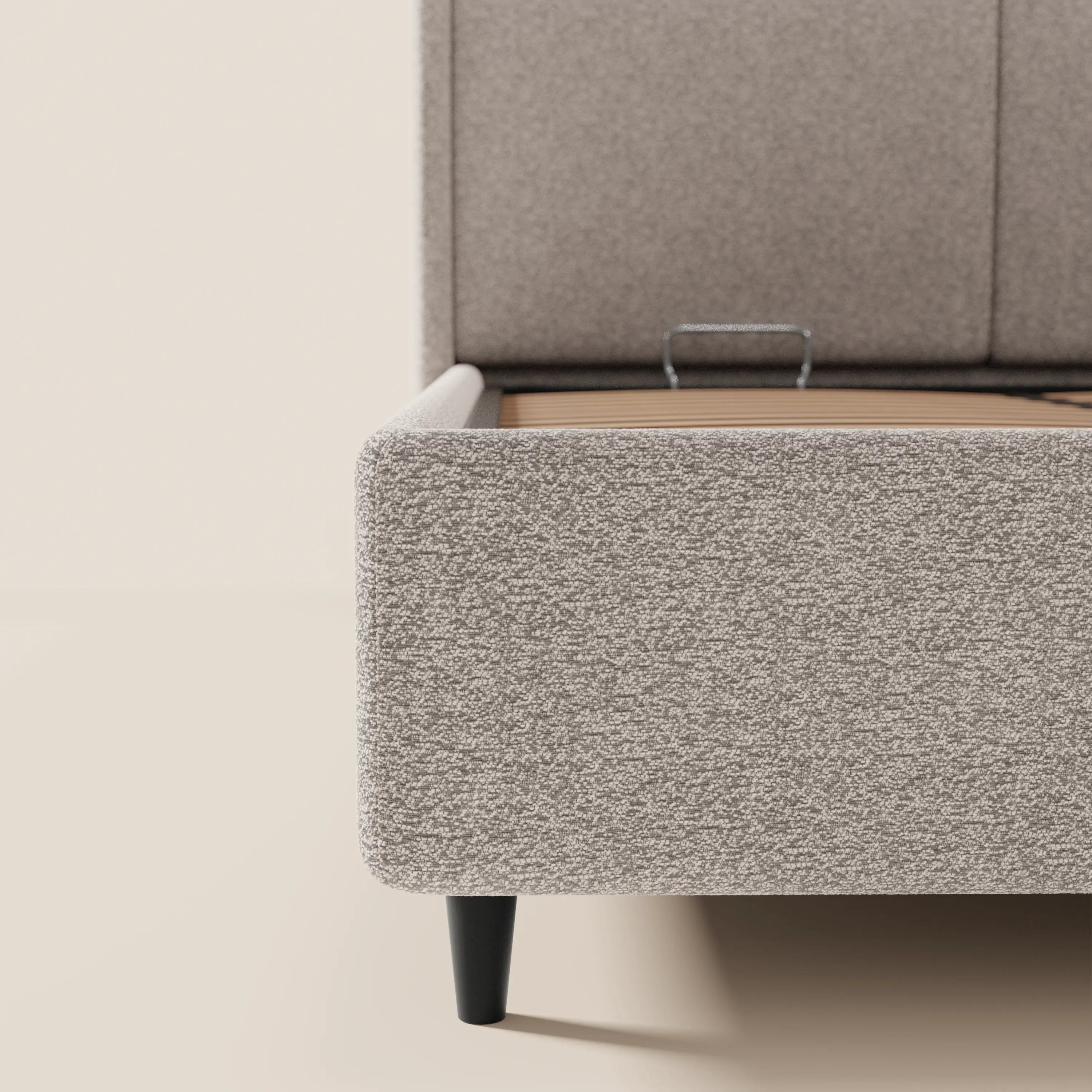 Un primo piano del letto di design VALKIRIA di Letti.store mette in risalto il tessuto beige chiaro certificato Oeko-Tex®, la finitura dalla morbida trama, gli angoli arrotondati, le gambe affusolate nere e i due cuscini imbottiti su uno sfondo neutro. Tra le caratteristiche figurano un vano contenitore e una testiera avvolgente.