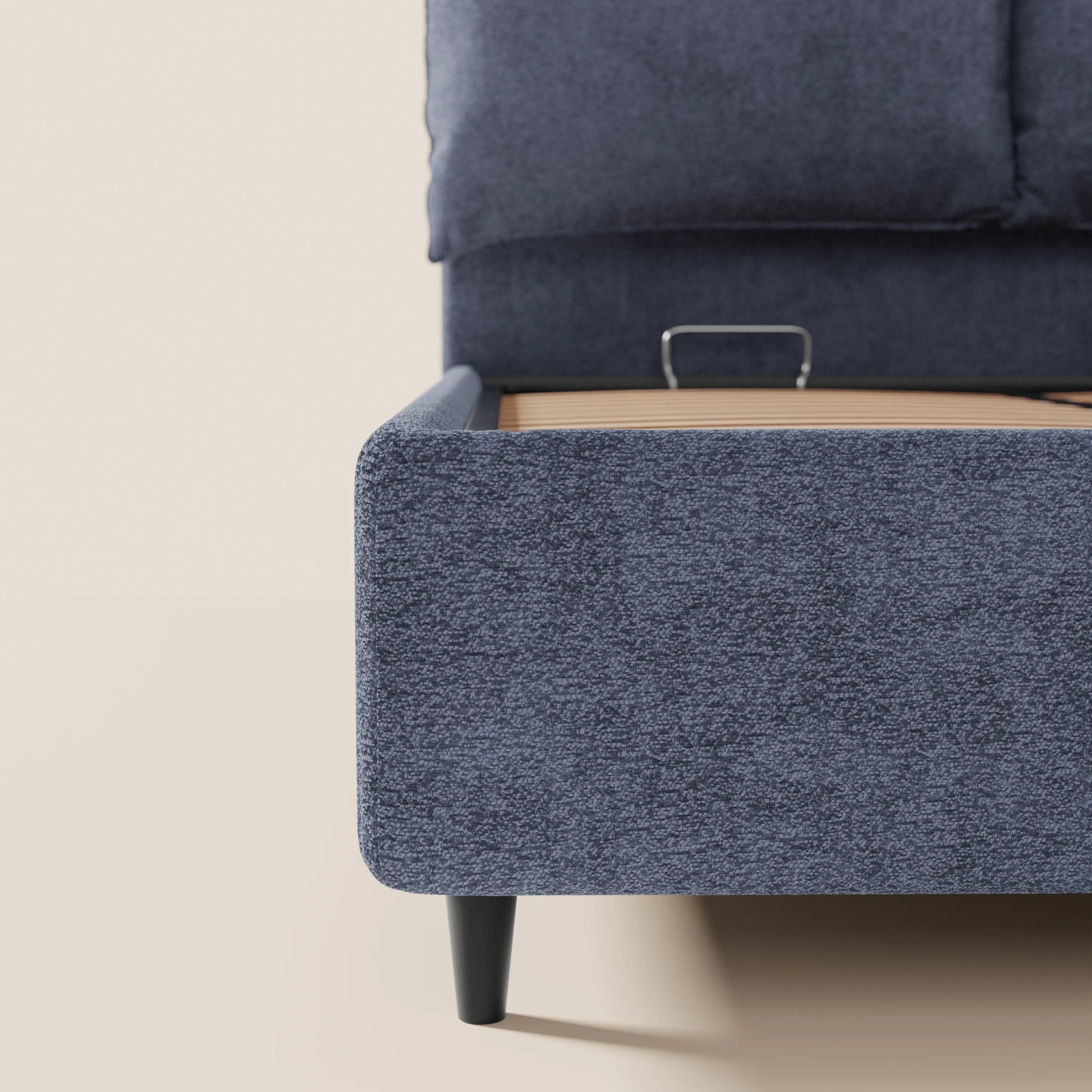 Primo piano del letto imbottito ZAYRON di Letti.store con vano contenitore, realizzato in tessuto Oeko-Tex® blu scuro con trama in rilievo, con angoli arrotondati, gambe affusolate nere, testiera a vista con cuscini regolabili e doghe in legno con staffa metallica. Sfondo bianco sporco.