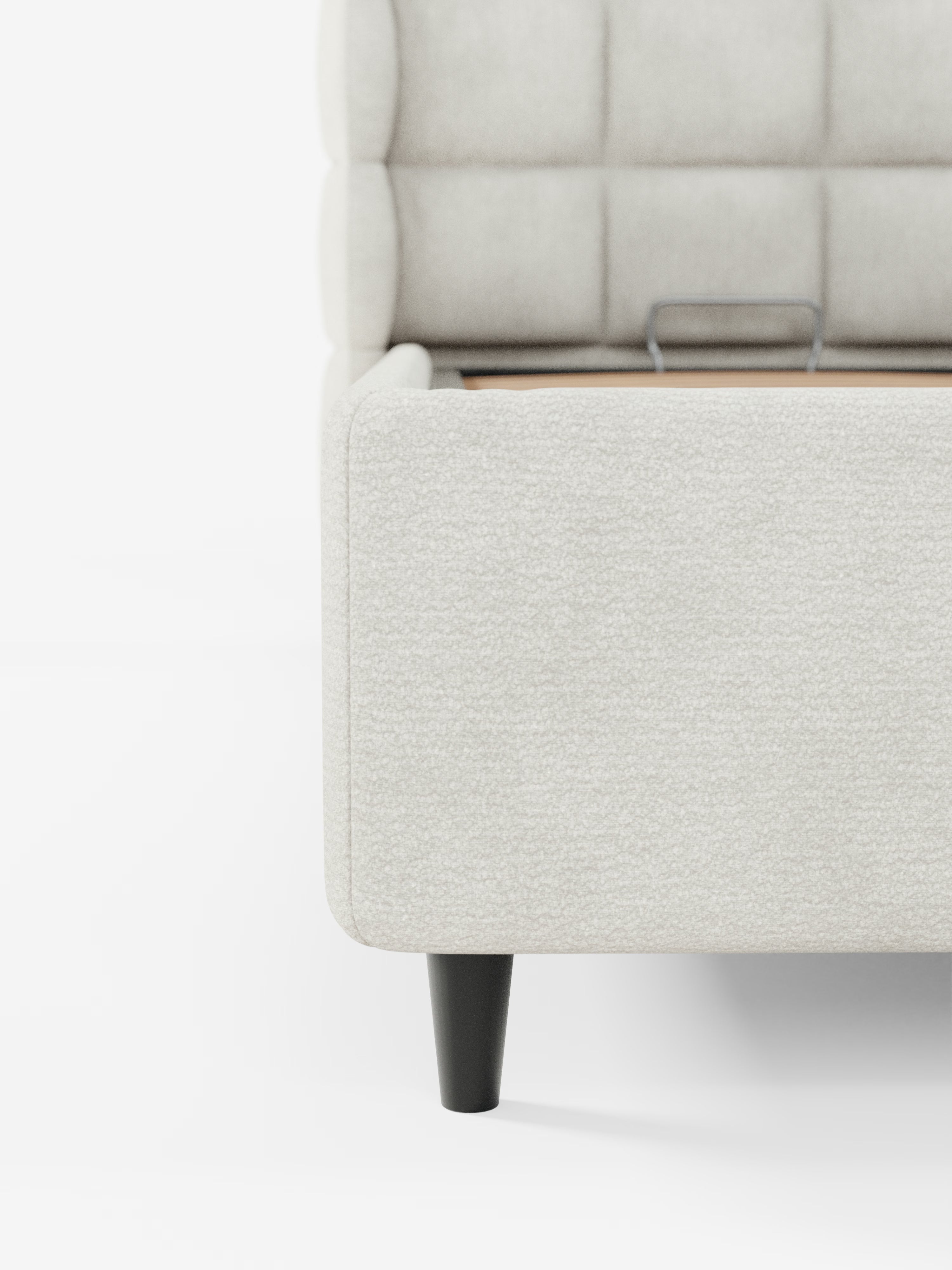 Primo piano dell'angolo del letto imbottito RAGNAR con contenitore e testiera avvolgente capitonné di Letti.store, realizzato in un elegante tessuto chiaro certificato Oeko-Tex®, con una gamba in legno scuro. Sono visibili anche la testiera con effetto capitonné e una maniglia in metallo sulla rete a doghe. Su uno sfondo bianco a tinta unita, questa foto mette in risalto l’eleganza moderna di questo complemento d’arredo per la camera da letto.