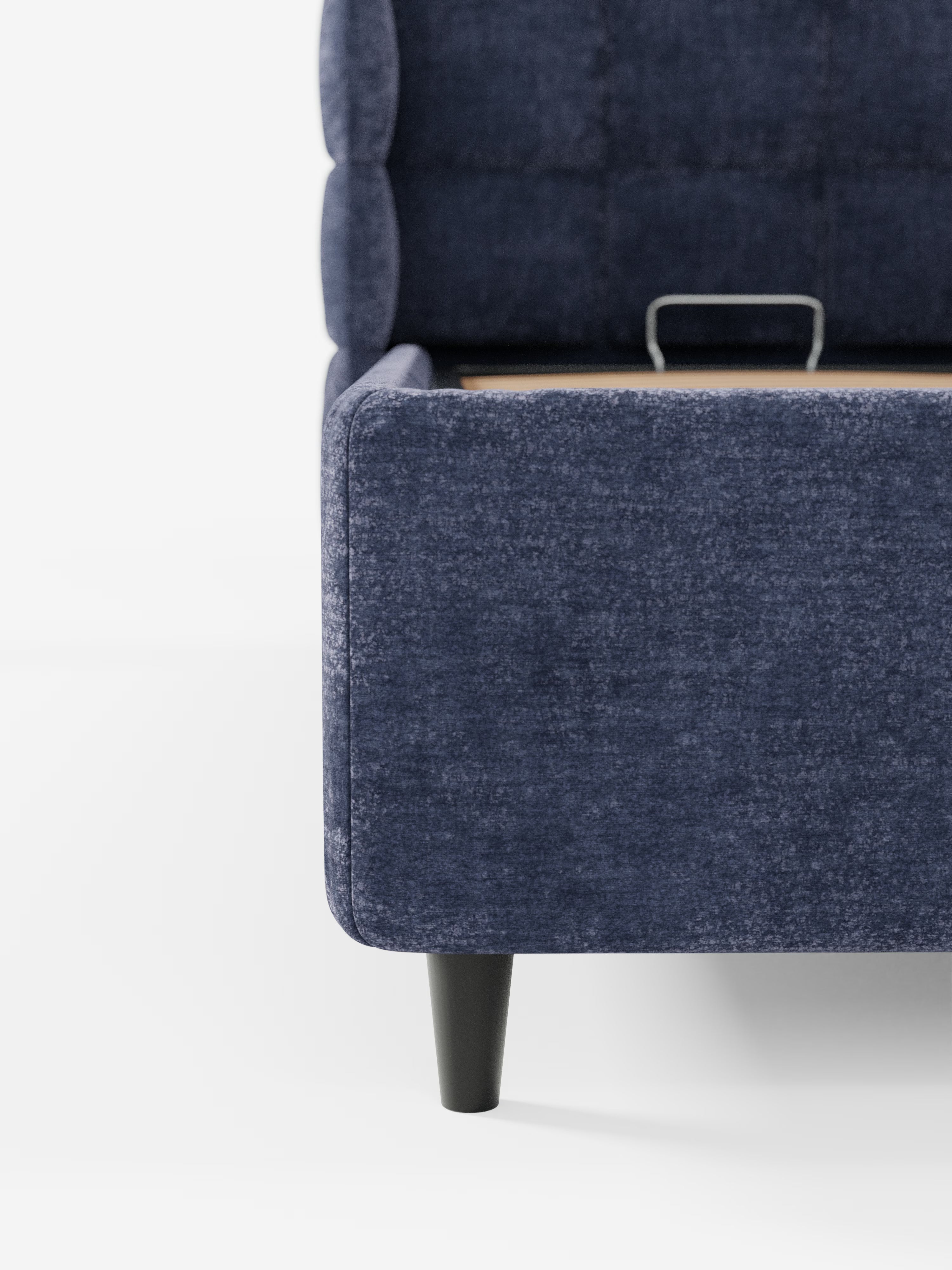 Primo piano del letto imbottito RAGNAR con contenitore e testiera capitonné avvolgente di Letti.store, rivestito in un elegante tessuto blu scuro certificato Oeko-Tex®. L’immagine mette in risalto l’angolo arrotondato della pediera, la gamba nera affusolata e una sezione del pannello laterale su sfondo bianco, evidenziando il design elegante e contemporaneo del letto e la sua texture raffinata.