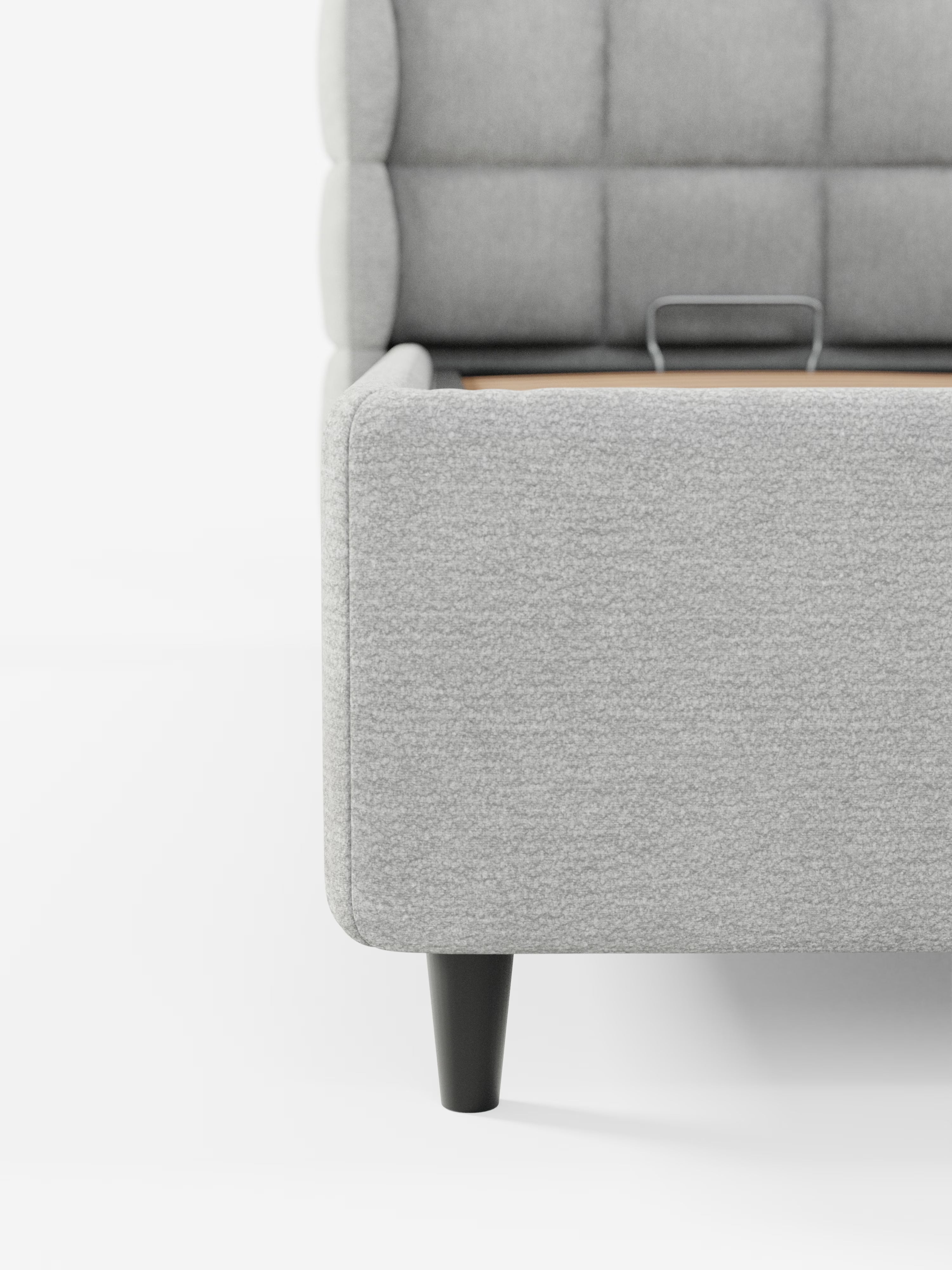 Primo piano dell'angolo inferiore del letto imbottito RAGNAR di Letti.store, dotato di vano contenitore e testiera capitonné avvolgente in tessuto grigio chiaro certificato Oeko-Tex®, con gambe nere. Il lato mostra un’elegante trapuntatura capitonné sulla testiera, mentre all’interno del telaio è visibile una piccola maniglia in metallo, che spicca su uno sfondo bianco a tinta unita.