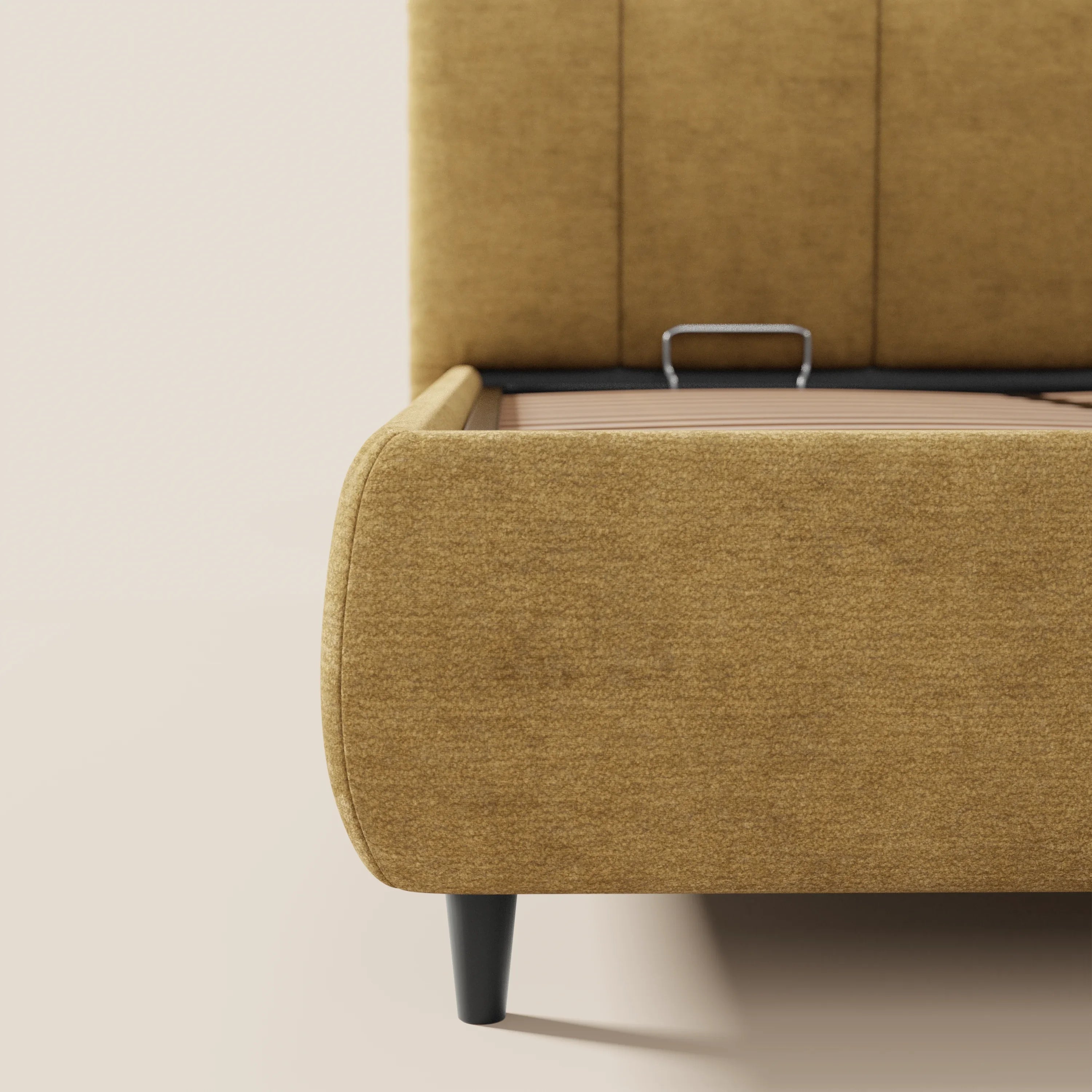 Primo piano di ARKAM di Letti.store, un letto imbottito color senape con vano contenitore e testiera a lavorazione verticale in tessuto Oeko-Tex®, dal design shabby chic, con gambe affusolate nere, che fa da contrasto con pareti e pavimento beige chiaro.