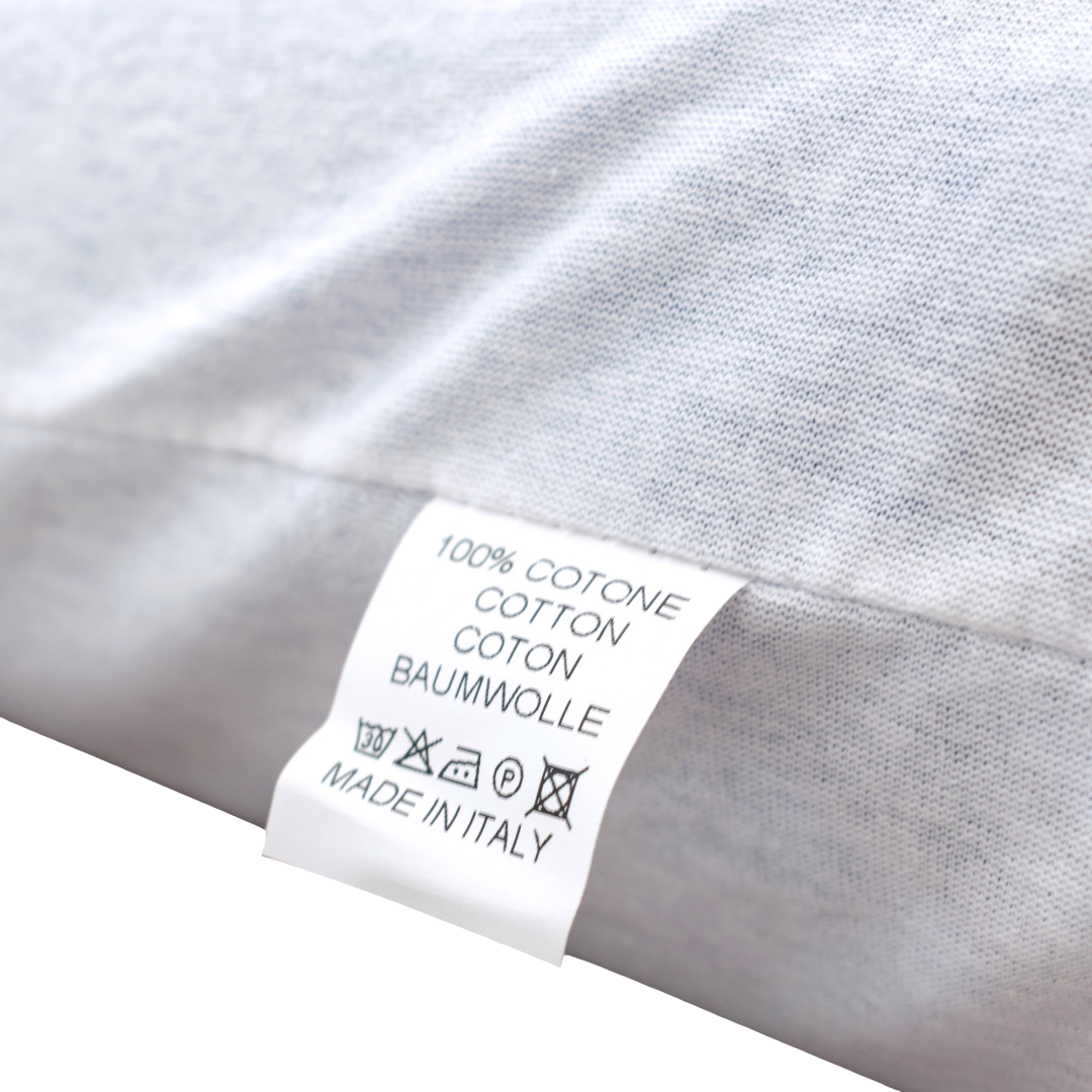 Un primo piano di un'etichetta in tessuto bianco su un capo tessile grigio chiaro mostra una scritta multilingue con le diciture “100% COTONE”, “COTTON”, “COTON” e “BAUMWOLLE”, insieme ai simboli di lavaggio e alla frase “MADE IN ITALY”. Cucita nella cucitura, questa etichetta attesta l’eccellente qualità del prodotto. L’articolo in questione è il Divani.Store AERION-BREEZE – Guanciale in Memory Gel rinfrescante e termoregolatore, dotato di avanzata tecnologia Polygel e Memory Gel per un supporto cervicale