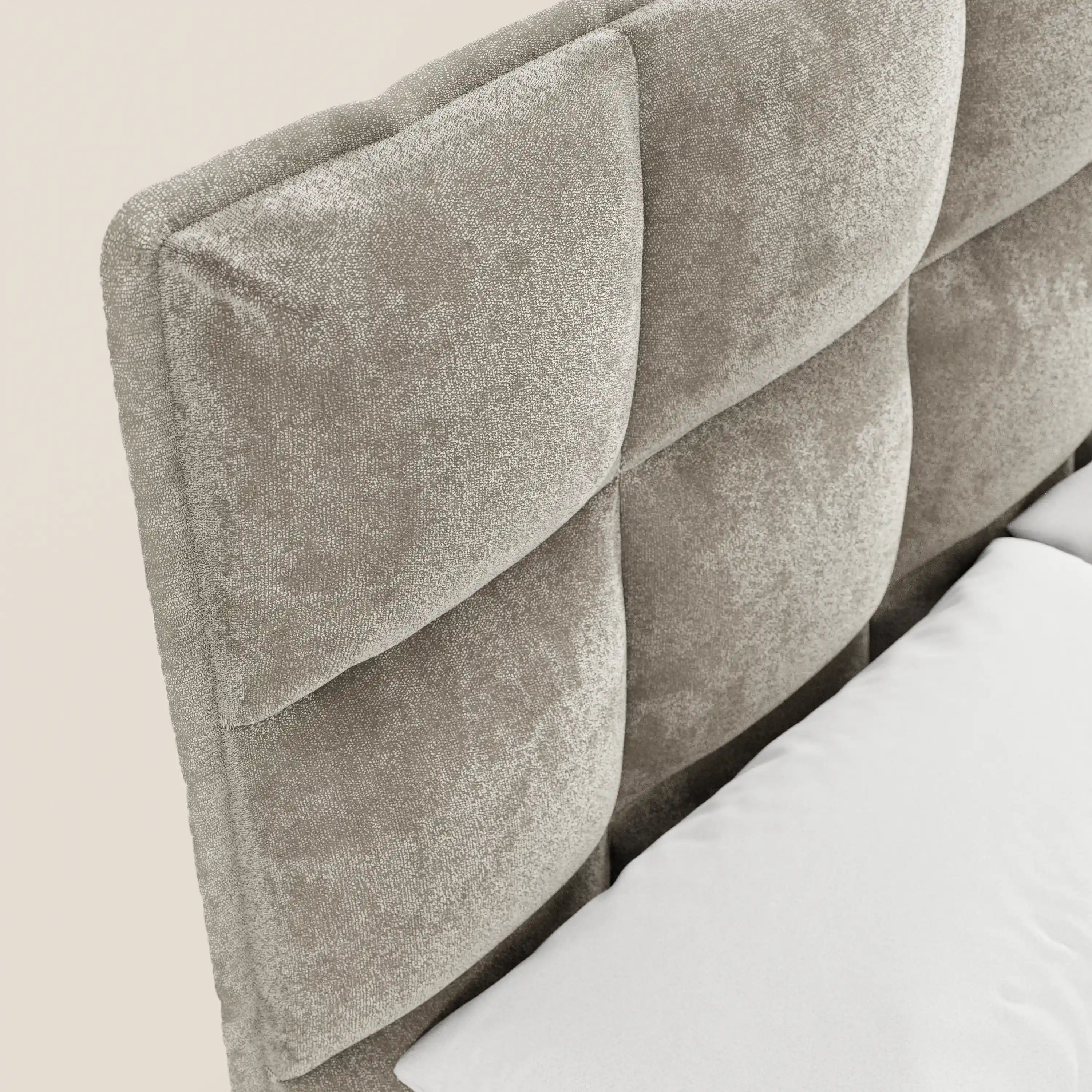 Primo piano del modello AURO di Letti.store: testiera capitonné alta di colore grigio chiaro, realizzata in morbida ciniglia certificata Oeko-Tex® con ampie sezioni trapuntate a quadrati. Include base contenitore; si notano cuscino e lenzuolo bianchi su sfondo bianco sporco.