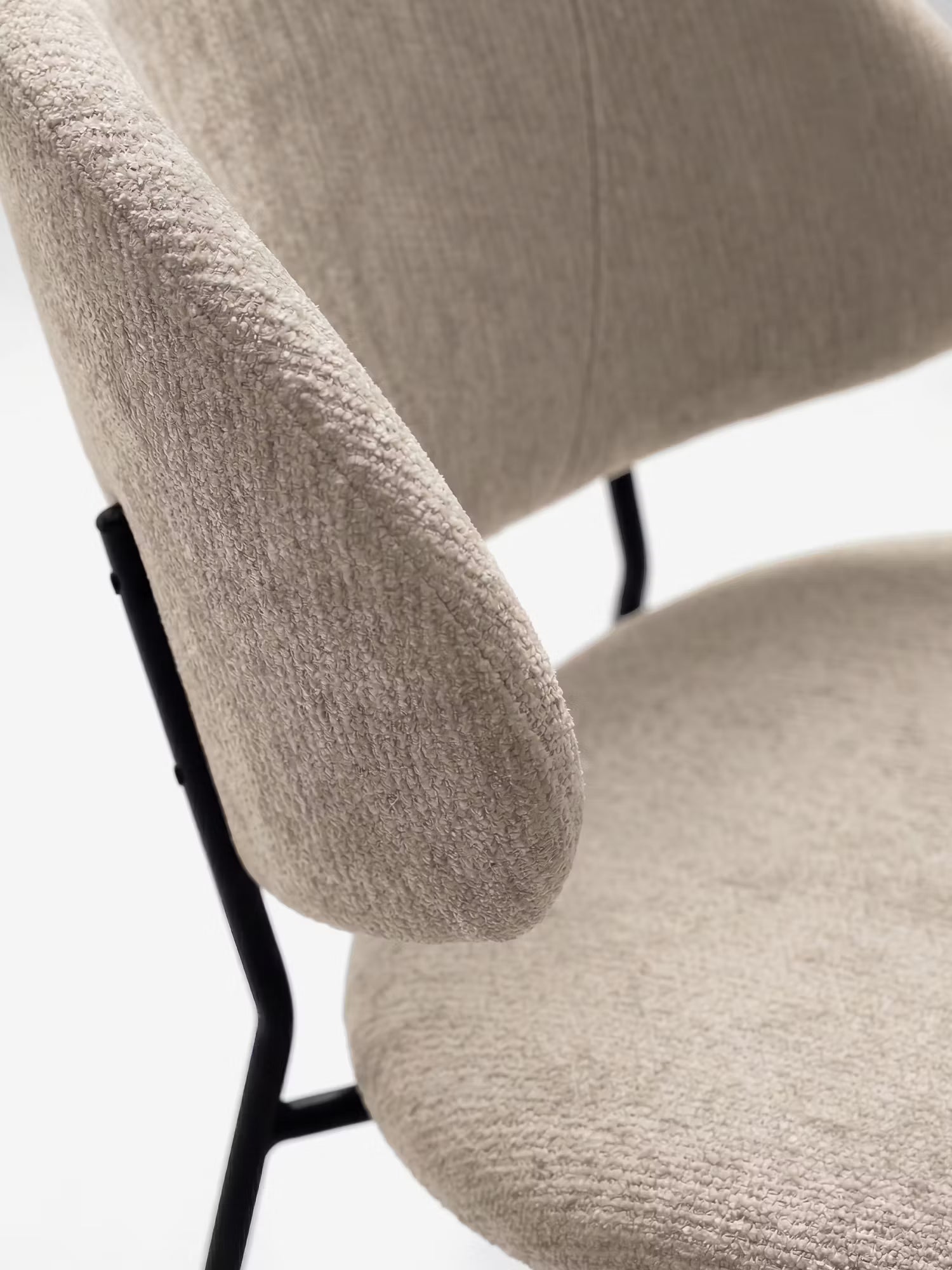 Primo piano della sedia Bruma di Divani.Store: dallo stile minimalista, con schienale avvolgente e seduta in elegante tessuto goffrato beige chiaro, oltre a sottili gambe in metallo nero. Design contemporaneo messo in risalto da uno sfondo chiaro e uniforme.