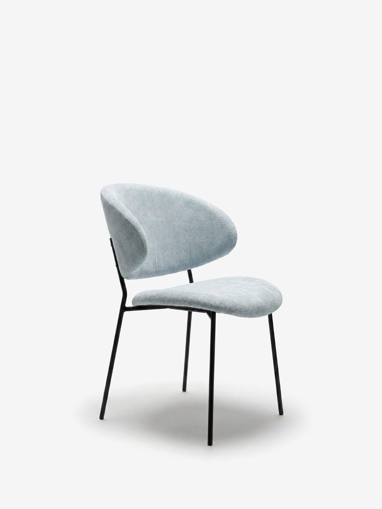 La sedia Bruma di Divani.Store è caratterizzata da un elegante tessuto goffrato e da uno schienale avvolgente, sostenuto da sottili gambe in metallo nero. Questo design minimalista spicca con il suo look contemporaneo su uno sfondo bianco a tinta unita.