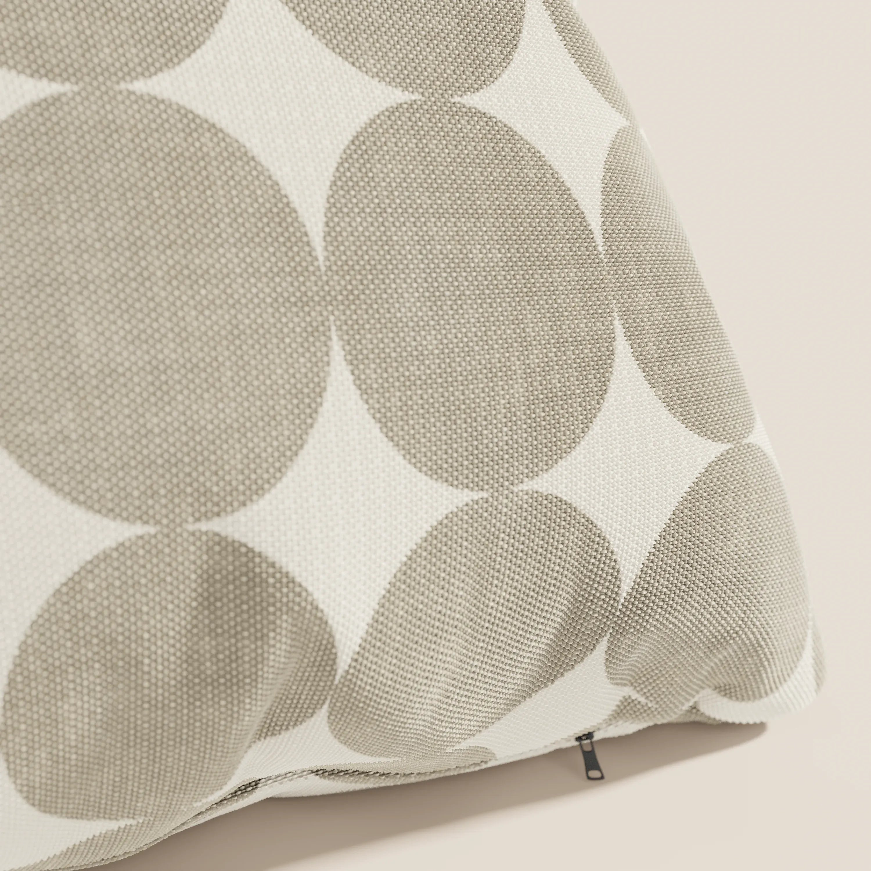 Un primo piano del Divani.Store Dot cuscini geometrici in tessuto idrorepellente beige mostra la sua copertina testurizzata con grandi cerchi sovrapposti di colore beige e bianco sporco su uno sfondo chiaro, oltre a una piccola chiusura lampo nera vicino al bordo inferiore.