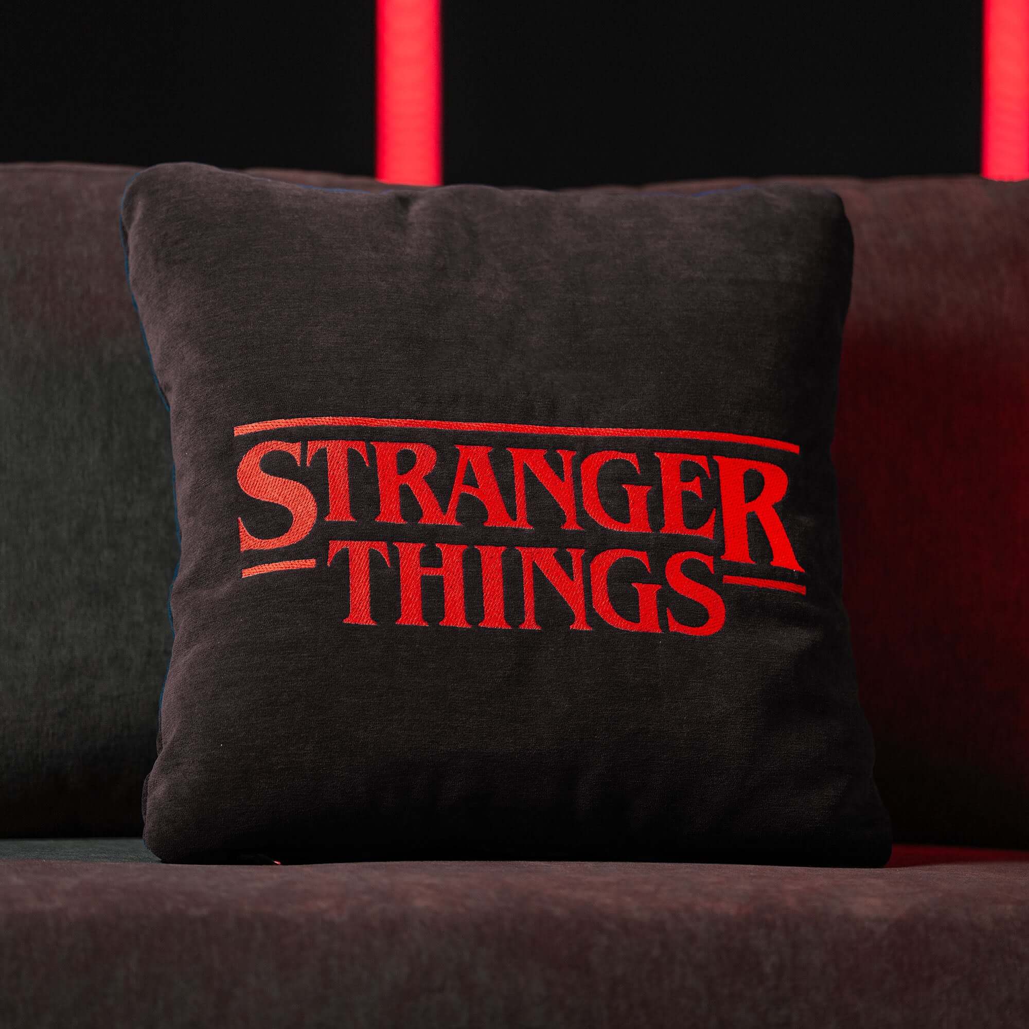 Un Cuscino Stranger Things Limited Edition con Ricamo 45x45 di Divani.Store, di colore antracite, è appoggiato su un divano scuro e mette in evidenza il logo rosso di "Stranger Things", con luci rosse verticali sfocate che creano uno sfondo drammatico.