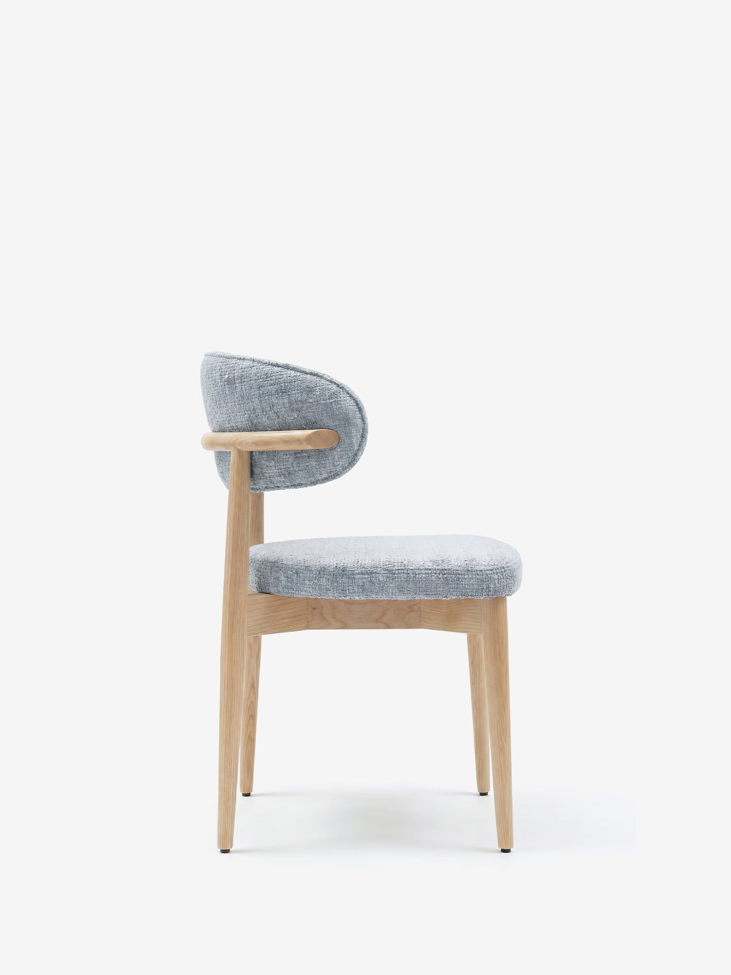 La sedia imbottita Dafne di Divani.Store presenta una linea minimalista con gambe e struttura in rovere naturale massiccio, oltre a un rivestimento in ciniglia certificato OEKO-TEX® in grigio chiaro, qui raffigurata di profilo su sfondo bianco.
