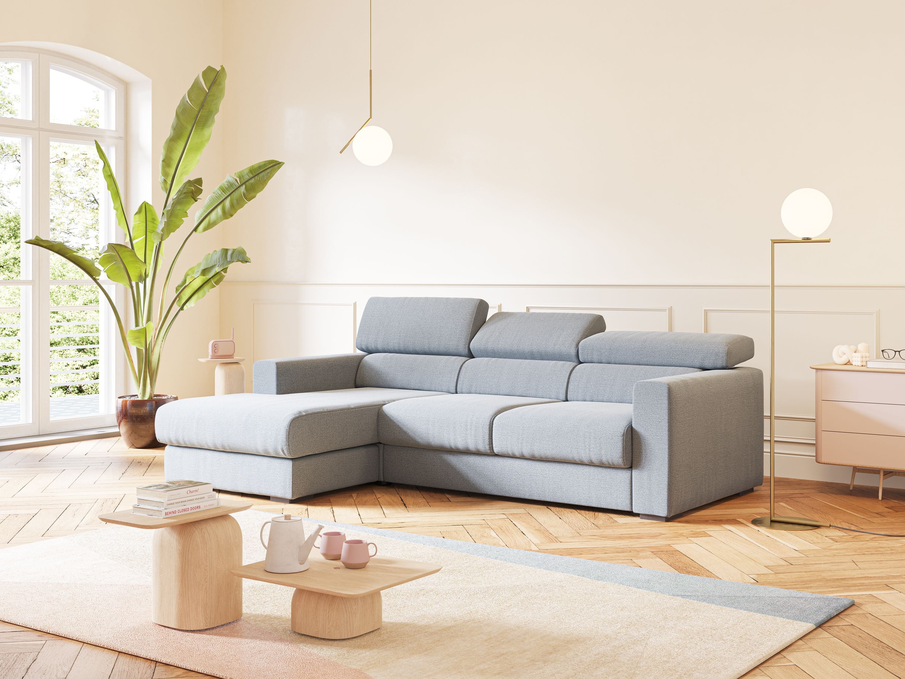 Un soggiorno moderno con il divano angolare Dylan con estraibile a cassettone in tessuto carta da zucchero di Divani.Store, pavimenti in legno, una grande pianta, lampade a sospensione e da terra rotonde, tavolini in legno chiaro con tazze e teiera, tappeto pastello e alte finestre illuminate dal sole.