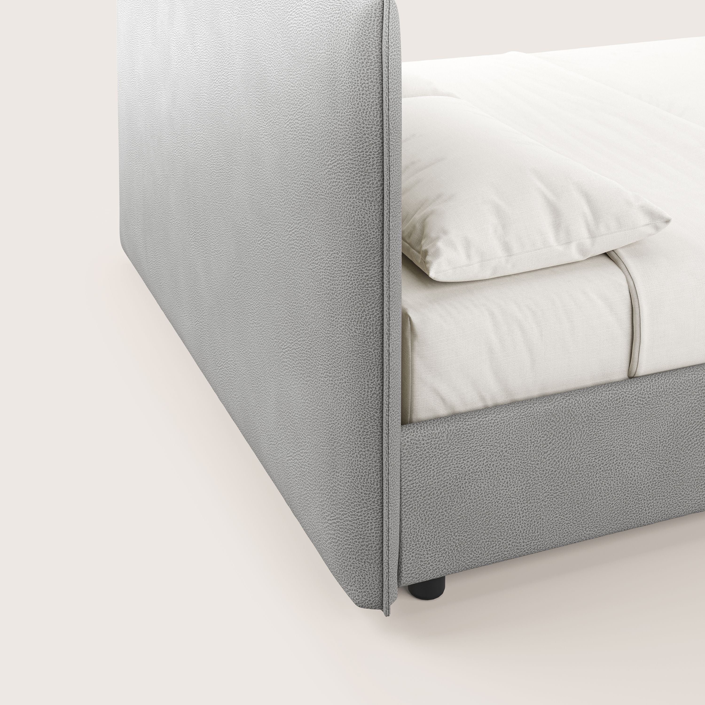 Primo piano di Nina letto contenitore di Letti.store: un letto dal design minimal con rivestimento in microfibra grigia antimacchia effetto pelle, con lenzuola e cuscino bianco sporco, visto di lato su uno sfondo neutro.