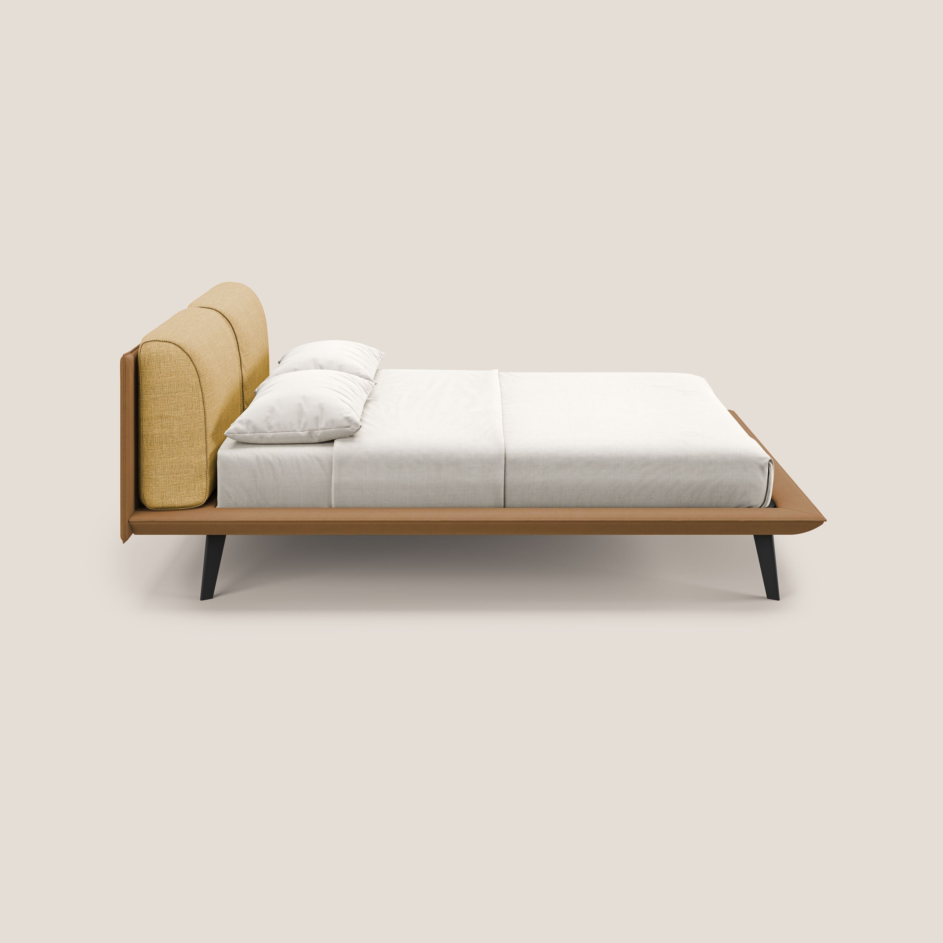 Il letto di design Petra di Letti.store è caratterizzato da un tessuto impermeabile bicolore (T11 cuoio, T06 giallo), cuscini in piuma, struttura in legno e gambe angolate in mogano. Mostrato in profilo laterale con biancheria da letto bianca e due cuscini su sfondo beige.