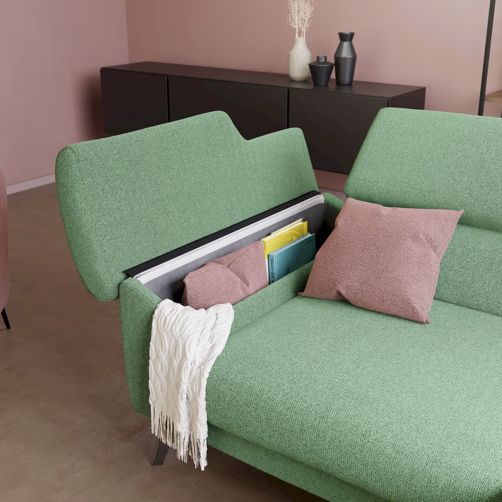 Il divano Donatello di Divani.Store in T23 menta ha sedute allungabili, braccioli contenitori e poggiatesta reclinabili. In una stanza minimalista dalle pareti rosa, il divano ospita libri e cuscini; una coperta color crema è drappeggiata sul lato vicino a un mobile marrone e a vasi.