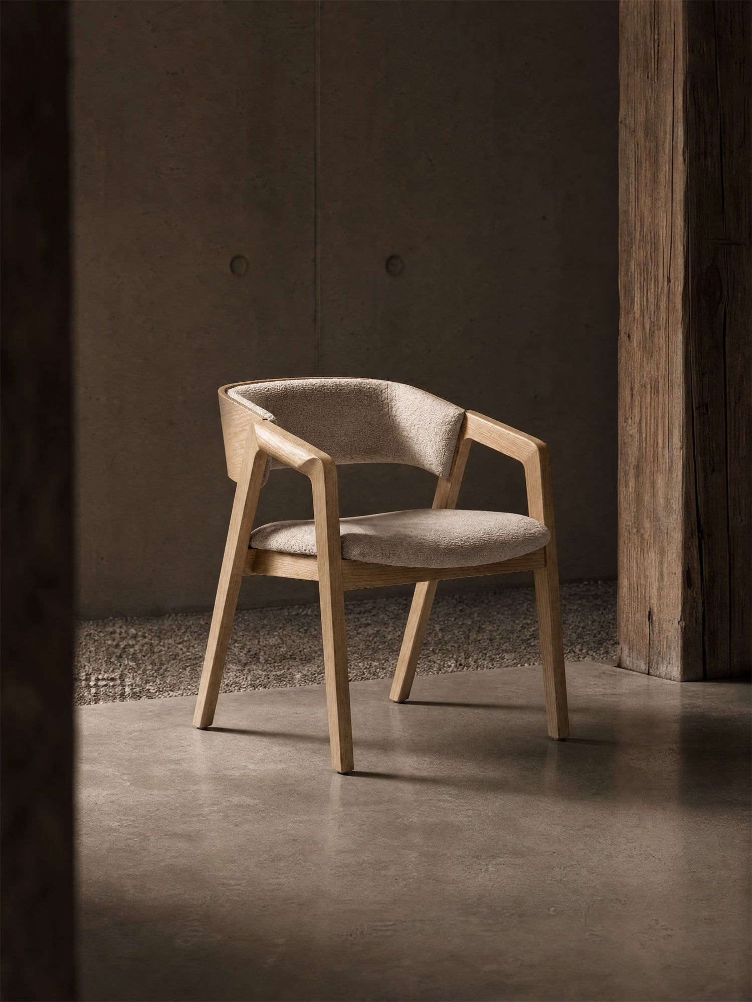 La sedia Etra in legno massello naturale con braccioli e rivestimento in tessuto OEKO-TEX® di Divani.Store è collocata su un pavimento in cemento levigato in una stanza dallo stile minimalista, dai toni terrosi, con una parete ruvida e un pilastro rustico in legno, illuminata da luci soffuse che creano ombre delicate.