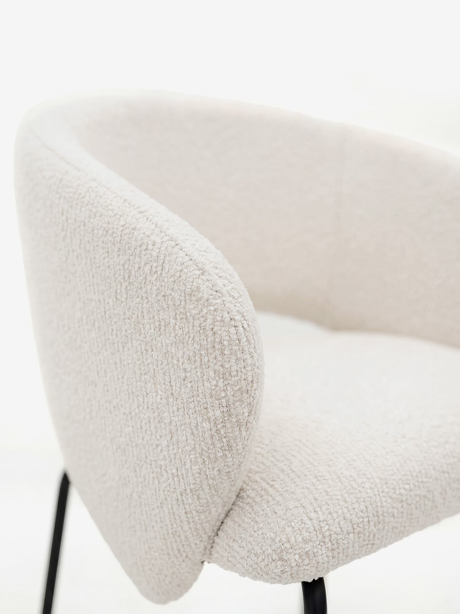 Un primo piano della sedia imbottita Fosca di Divani.Store, con braccioli di design in morbido tessuto certificato OEKO-TEX® e gambe sottili in metallo nero, dal rivestimento testurizzato e dalle linee curve su uno sfondo luminoso.