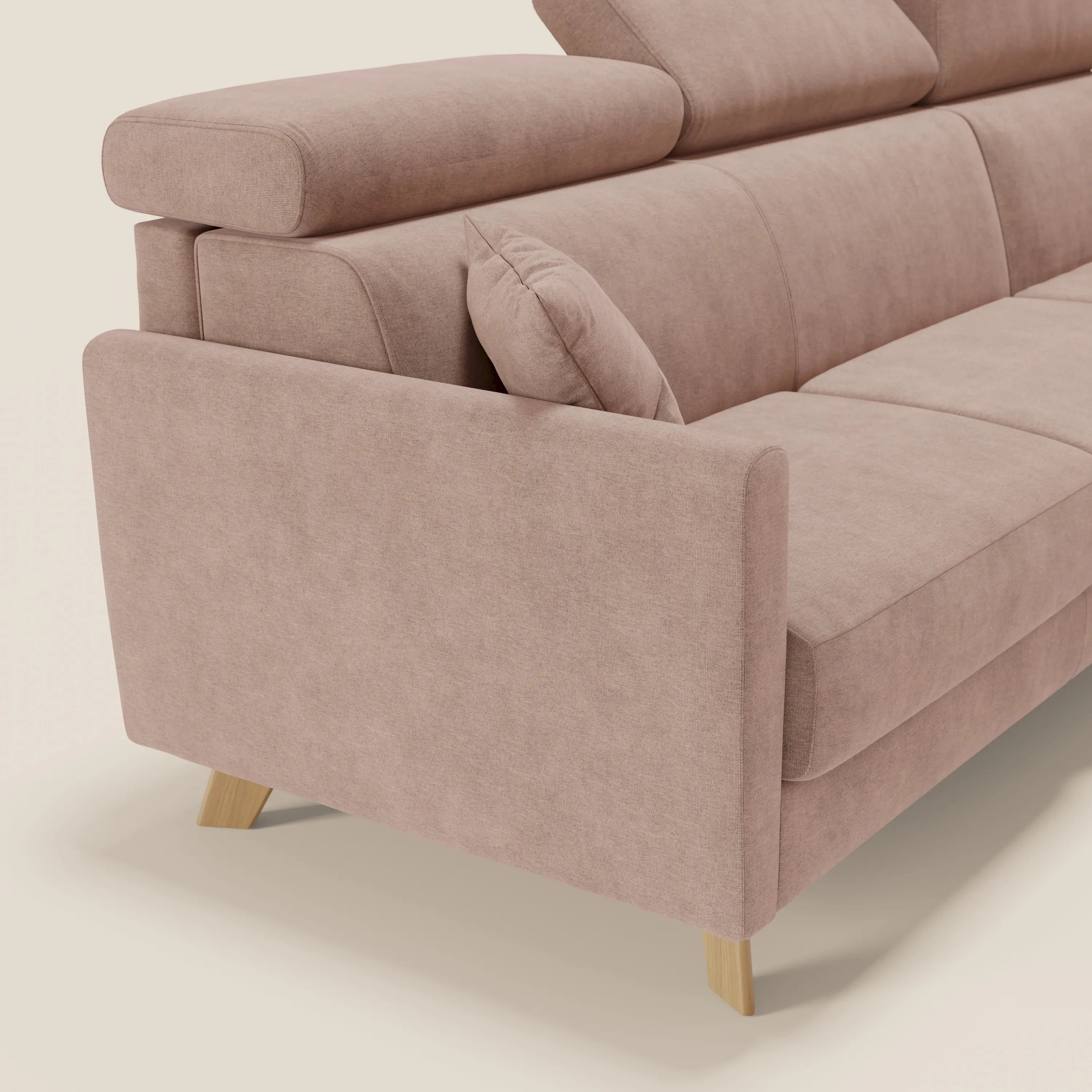 Primo piano del divano letto angolare Francy sfoderabile di Divani.Store in tessuto smacchiabile T17 rosa, con poggiatesta reclinabile, gambe in legno, cuscino d'accento accanto al bracciolo e sfondo bianco sporco.