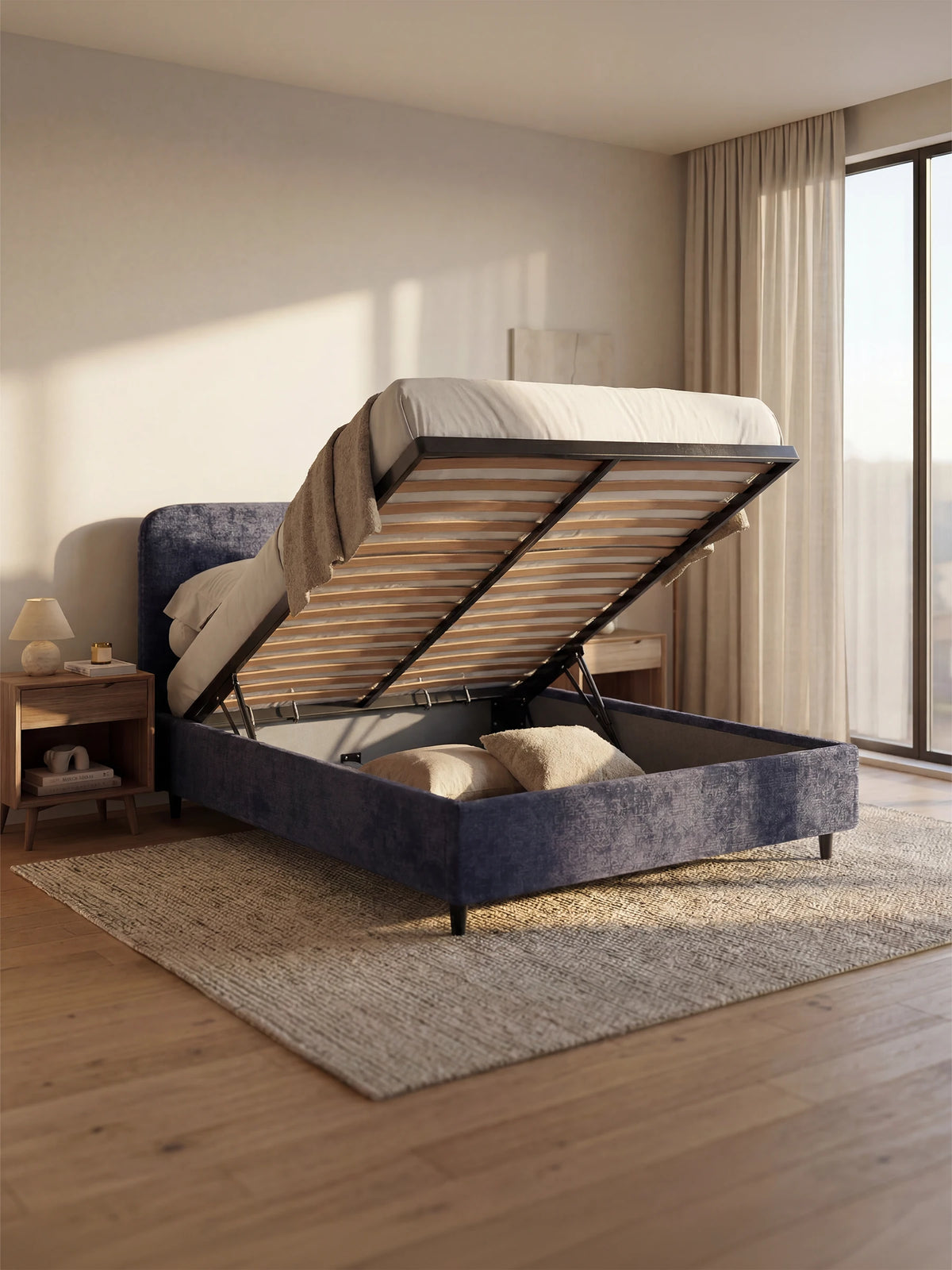 Un letto KAELOS blu, modello minimal imbottito – con vano contenitore, piedini alti e testiera arrotondata in tessuto ciniglia – è raffigurato con la base sollevata per mostrare lo spazio contenitivo, appoggiato su un tappeto testurizzato in una camera da letto minimalista.