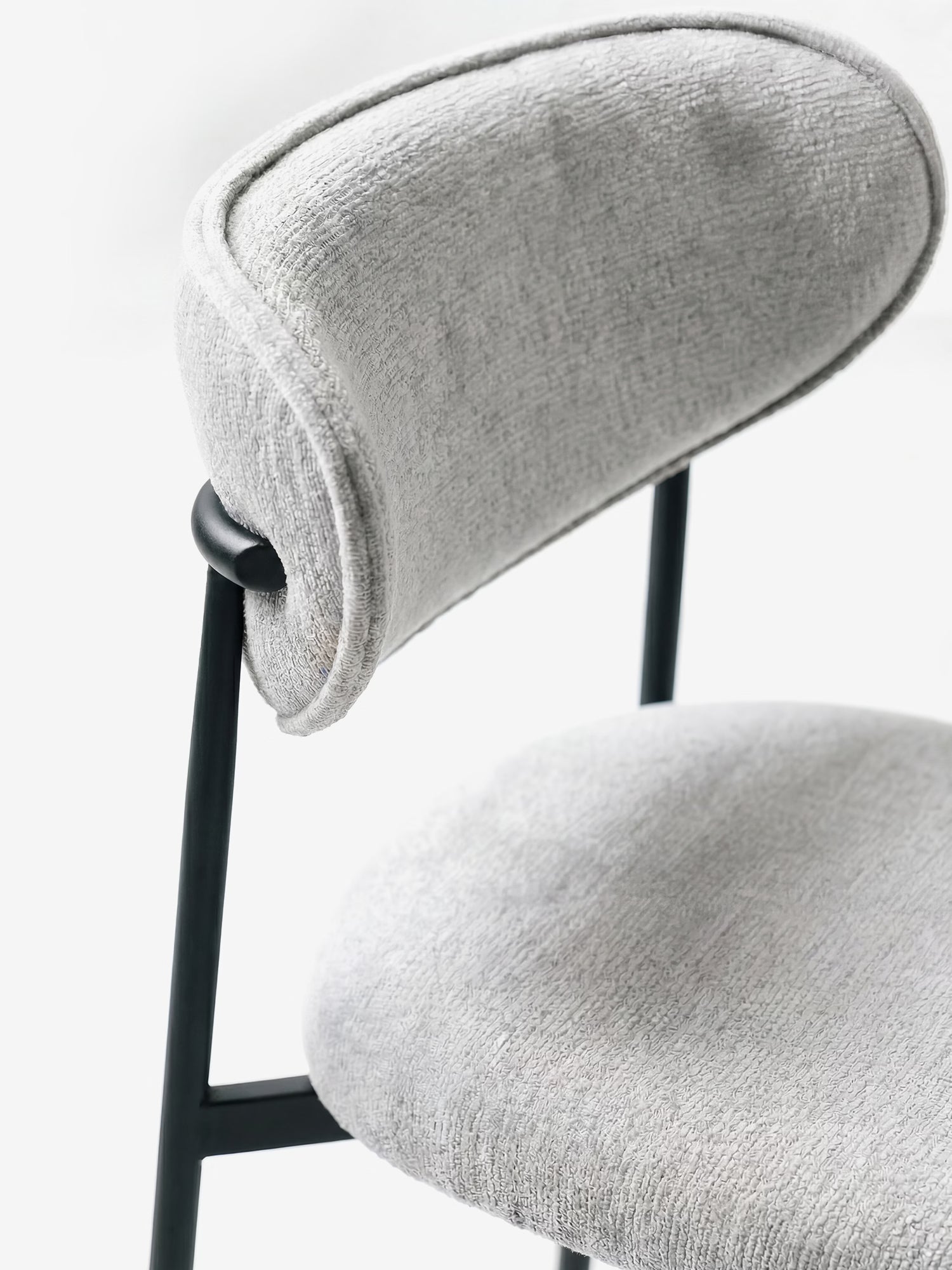 Un primo piano della Luce Sedia di Divani.Store, rivestita in morbido tessuto ciniglia chiaro, con schienale curvo e struttura in metallo nero, che mette in risalto il suo design moderno su uno sfondo bianco.