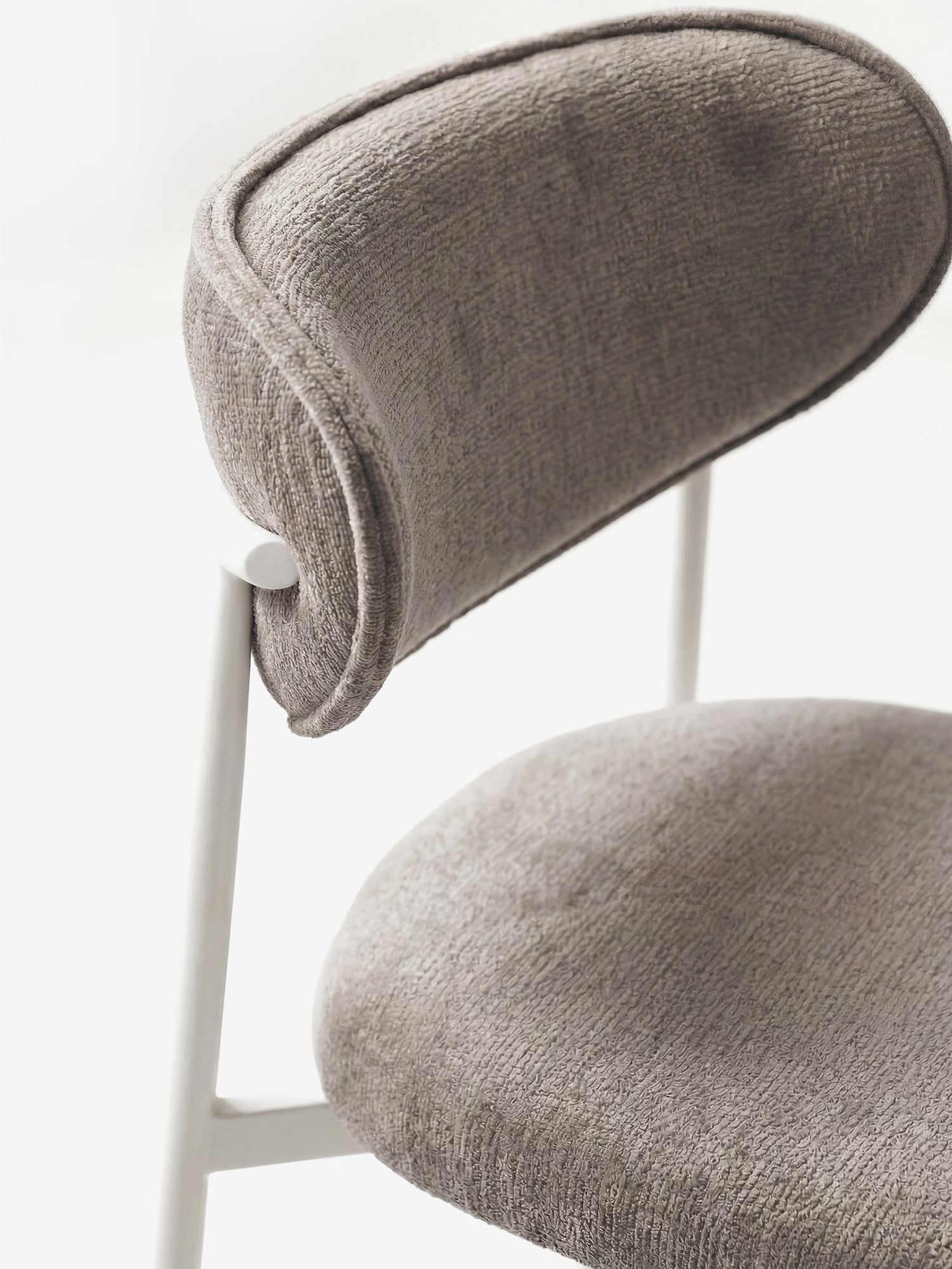 Primo piano della sedia imbottita “Luce” di Divani.Store, caratterizzata da schienale e seduta imbottiti e sinuosi, rivestiti in morbido tessuto grigio testurizzato, con una struttura minimalista in metallo bianco, su uno sfondo luminoso e sfocato.