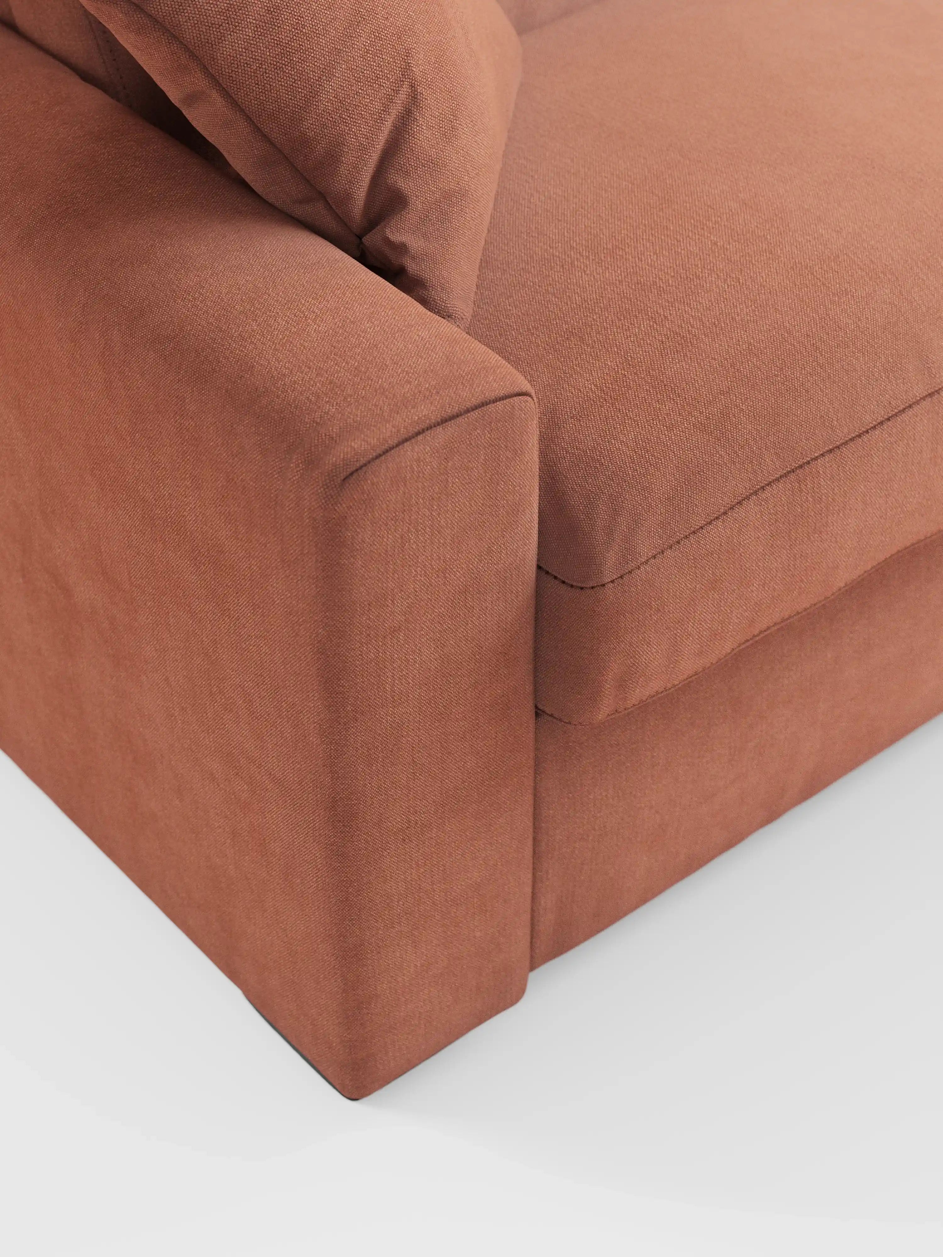 Primo piano di un divano letto Maximus a 3 posti con rivestimento in tessuto marrone e materasso ALTO da 20 cm di Divani.Store, con particolare attenzione al bracciolo e al cuscino di seduta, caratterizzati da cuciture pulite e da un tessuto Oeko-Tex morbido e resistente alle macchie. Lo sfondo bianco a tinta unita mette in risalto le linee moderne e squadrate del divano letto, la sua solida struttura e i poggiatesta regolabili.